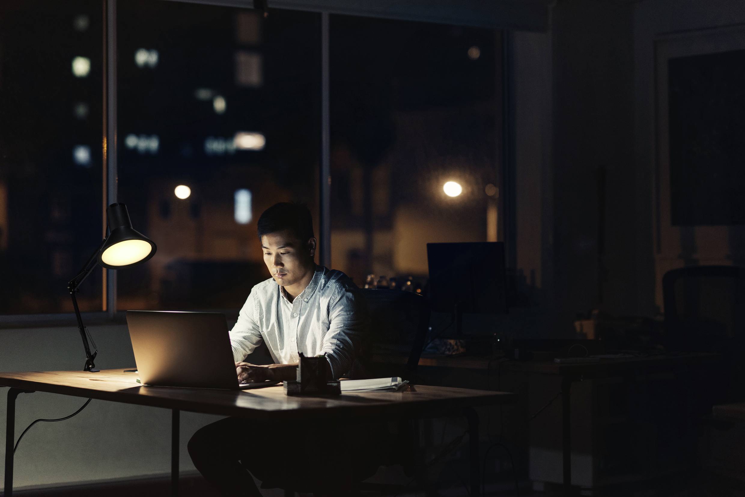 Osoba pracuje w nocy na laptopie w oświetlonym biurze, otoczona ciemnością i refleksami świateł z zewnątrz.
