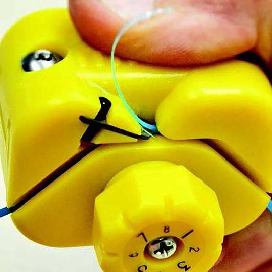 Zbliżenie żółtego urządzenia trzymanego w ręku, które napina drut. Ostrość skierowana na numerowanym pokrętle regulacyjnym i ułożeniu palców.