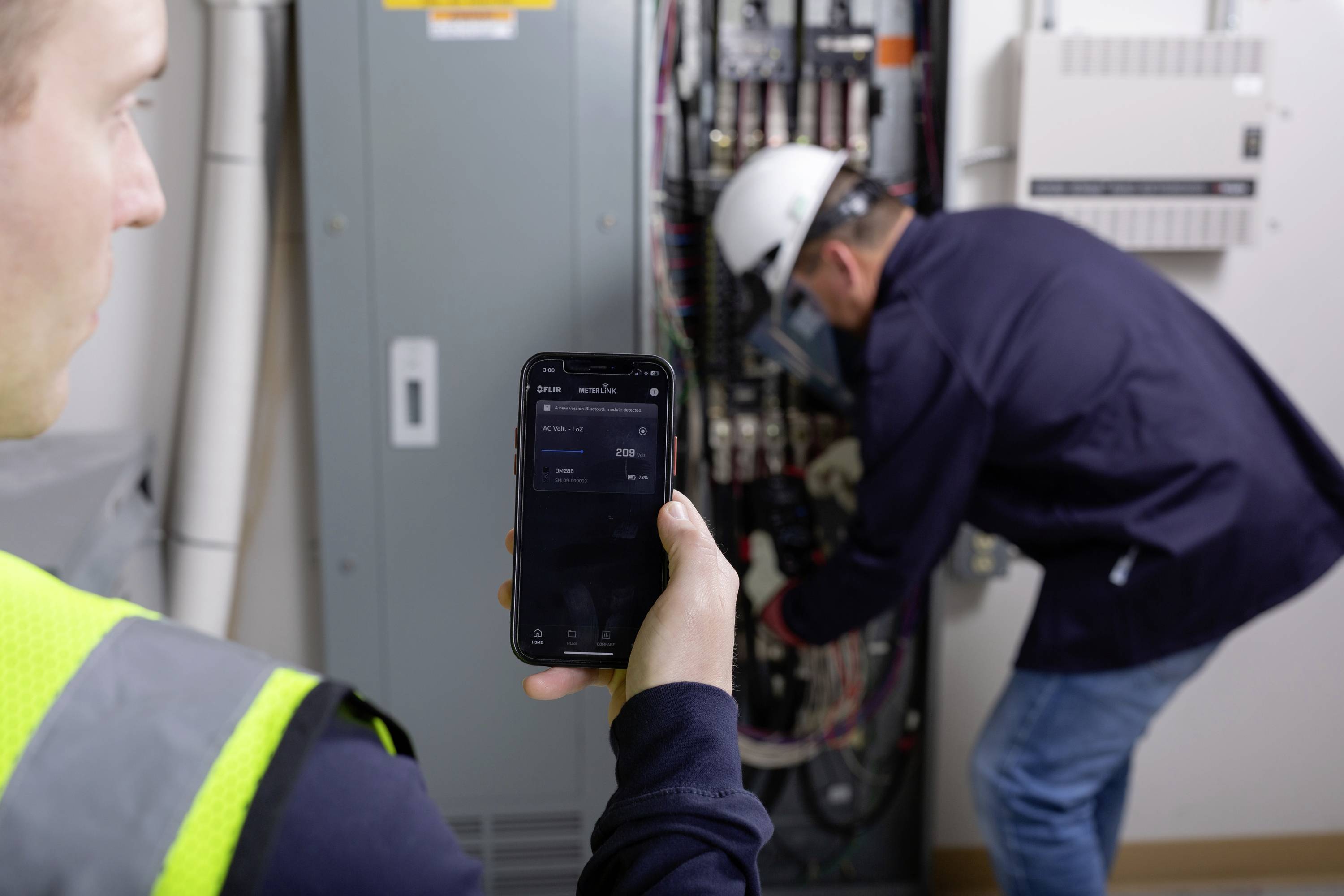 Robotnik stoi przed otwartą rozdzielnicą i używa smartfona do sprawdzenia zużycia energii elektrycznej. W tle widać pracującego technika.