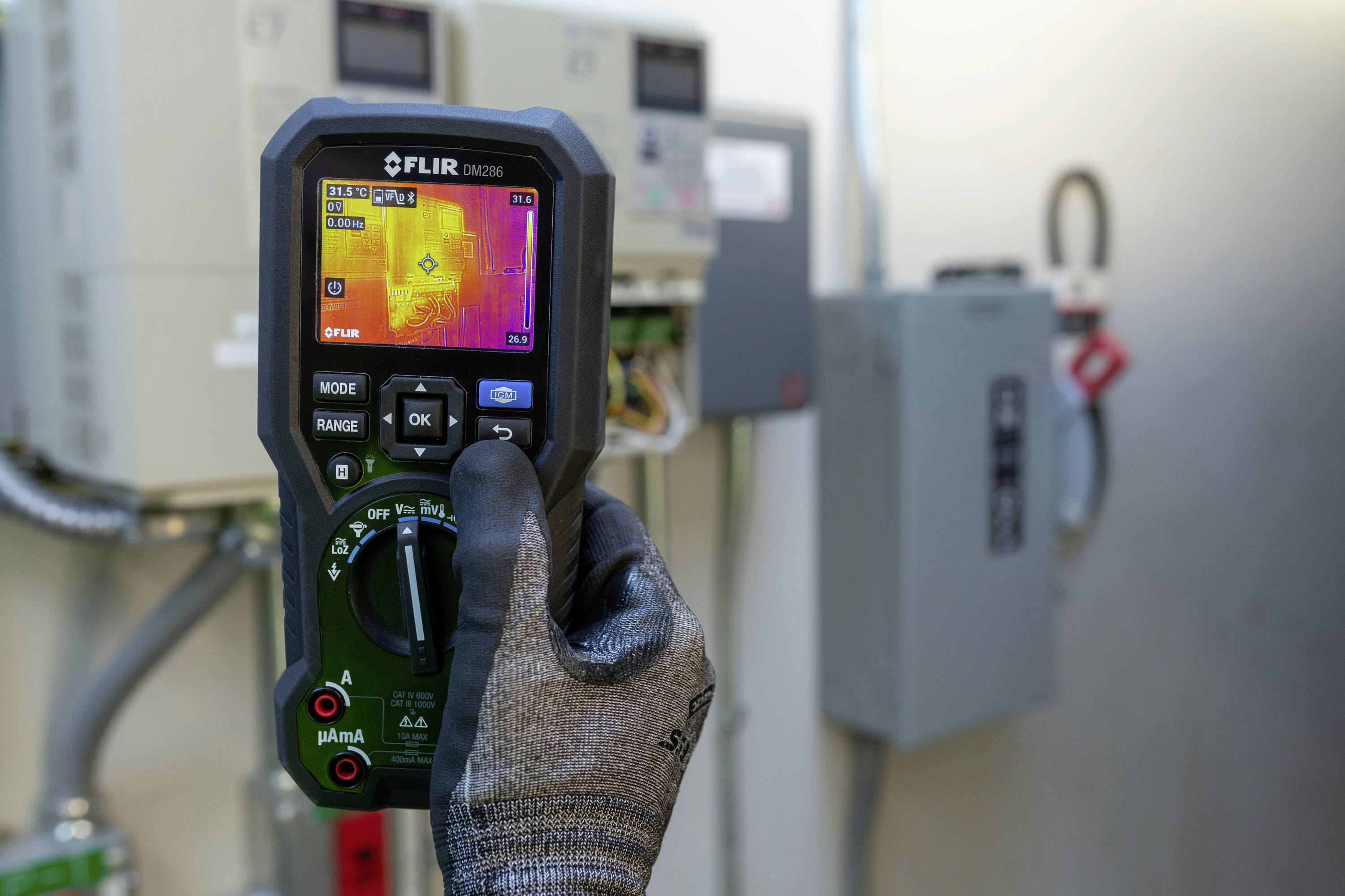 Osoba trzyma urządzenie kamery termowizyjnej przed szafkami elektrycznymi. Na ekranie widoczny jest rozkład temperatury.