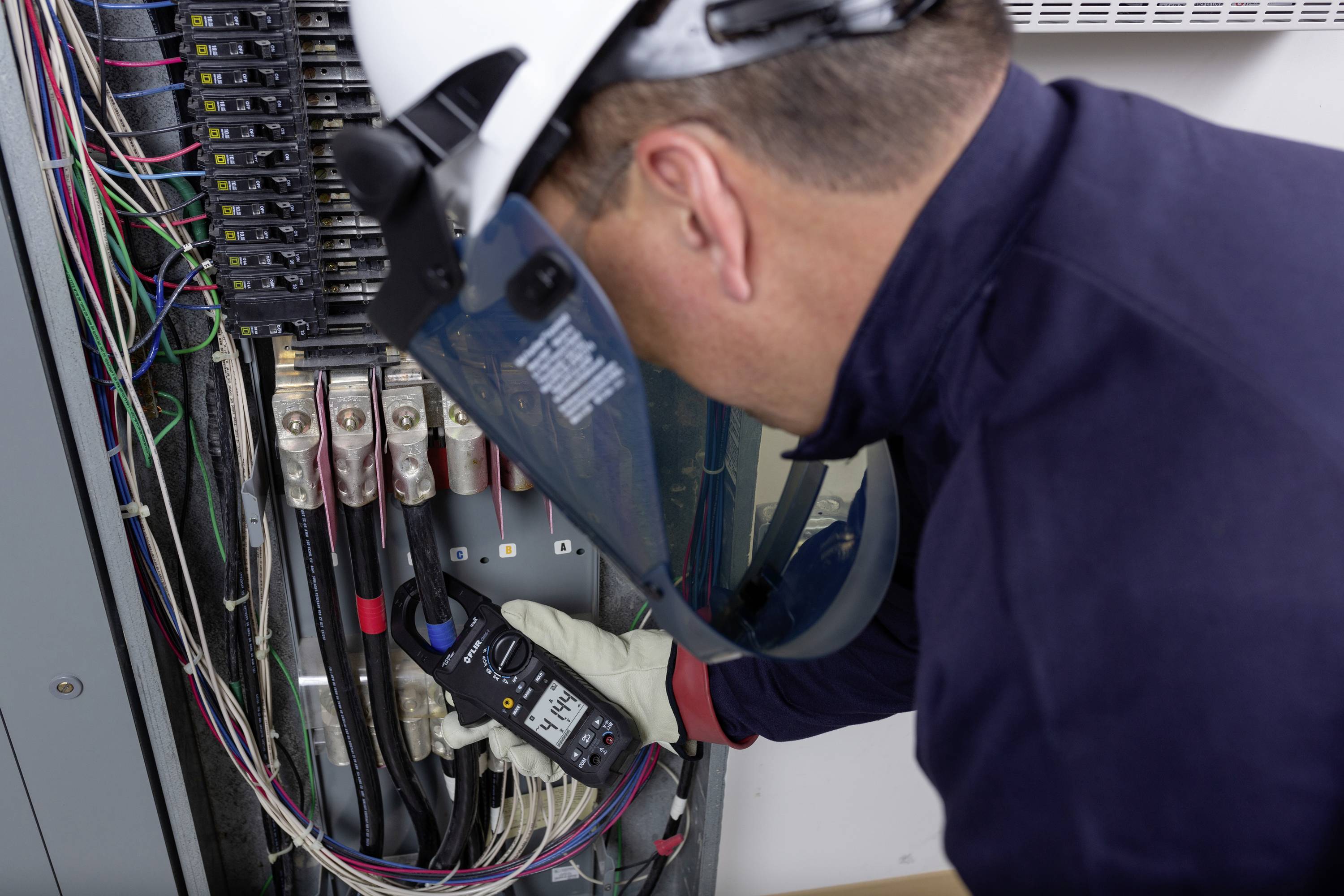 Elektryk w ubraniu ochronnym mierzy napięcie w szafce rozdzielczej za pomocą multimetru. Widocznych jest wiele kolorowych przewodów.
