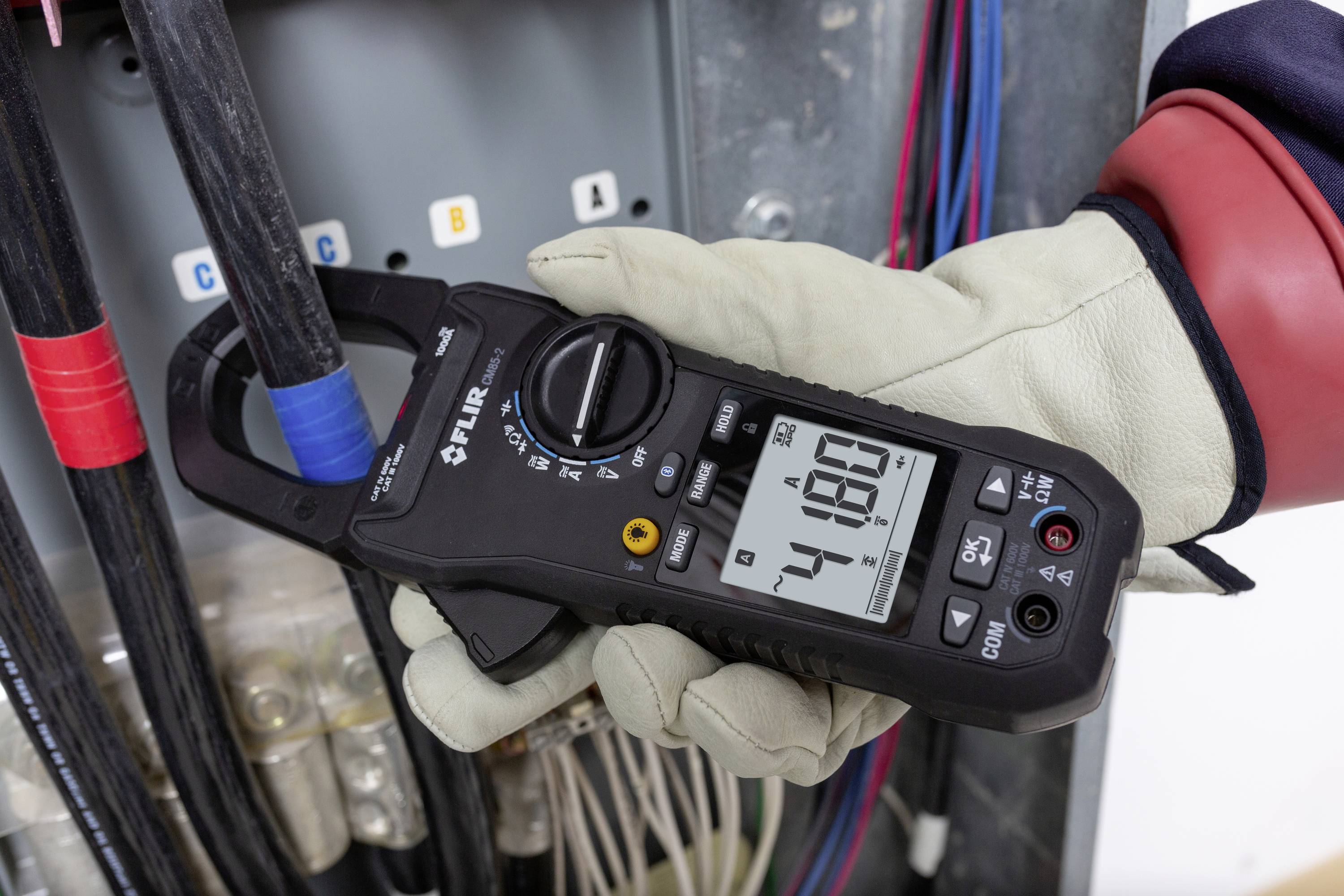 Dłoń w rękawicy trzyma cyfrowy multimetr wskazujący 48,0 amperów przed rozdzielnicą elektryczną.