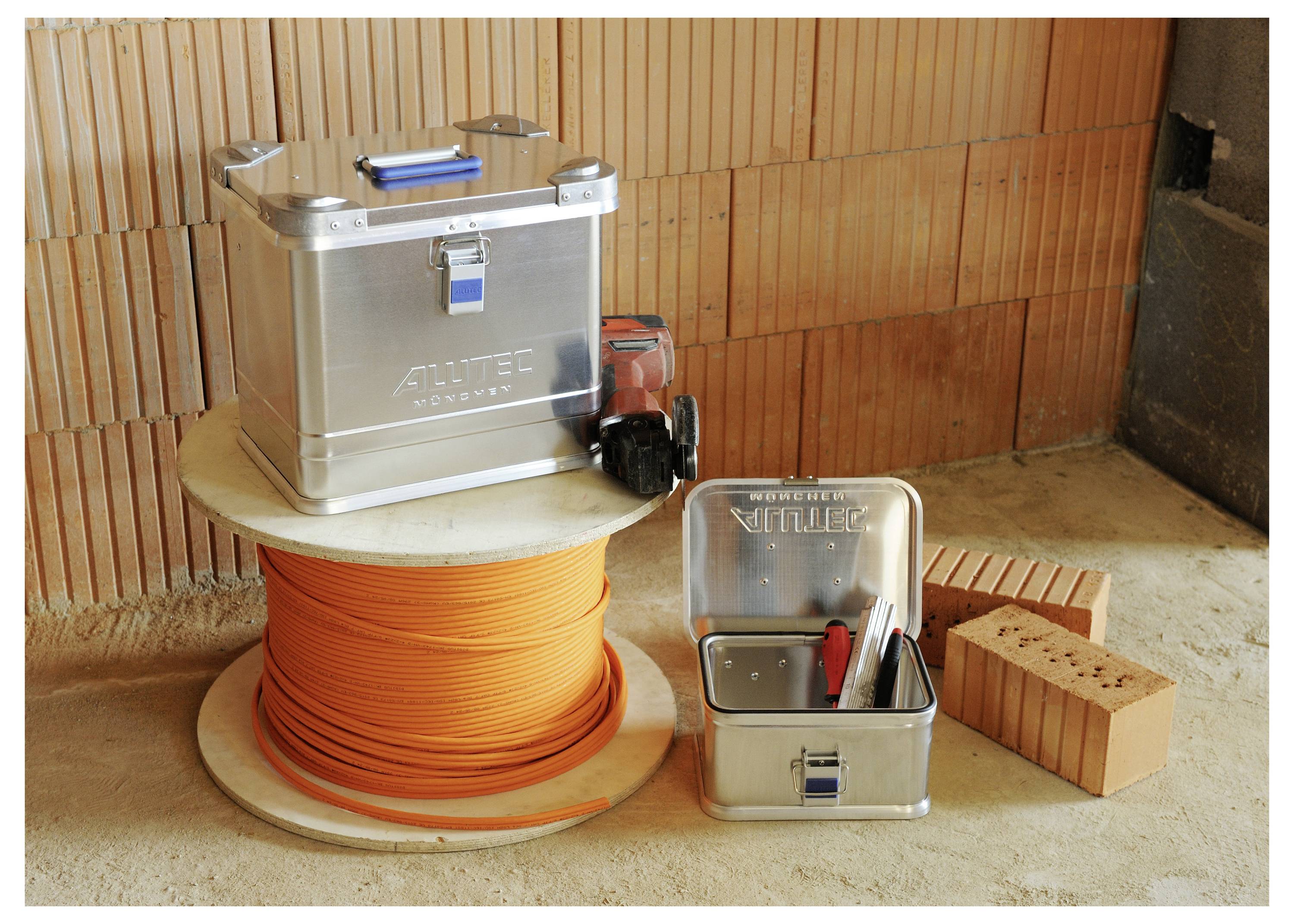 Szpula pomarańczowego przewodu elektrycznego i dwie metalowe skrzynie narzędziowe na placu budowy, z czerwoną wiertarką i cegłami w pobliżu.