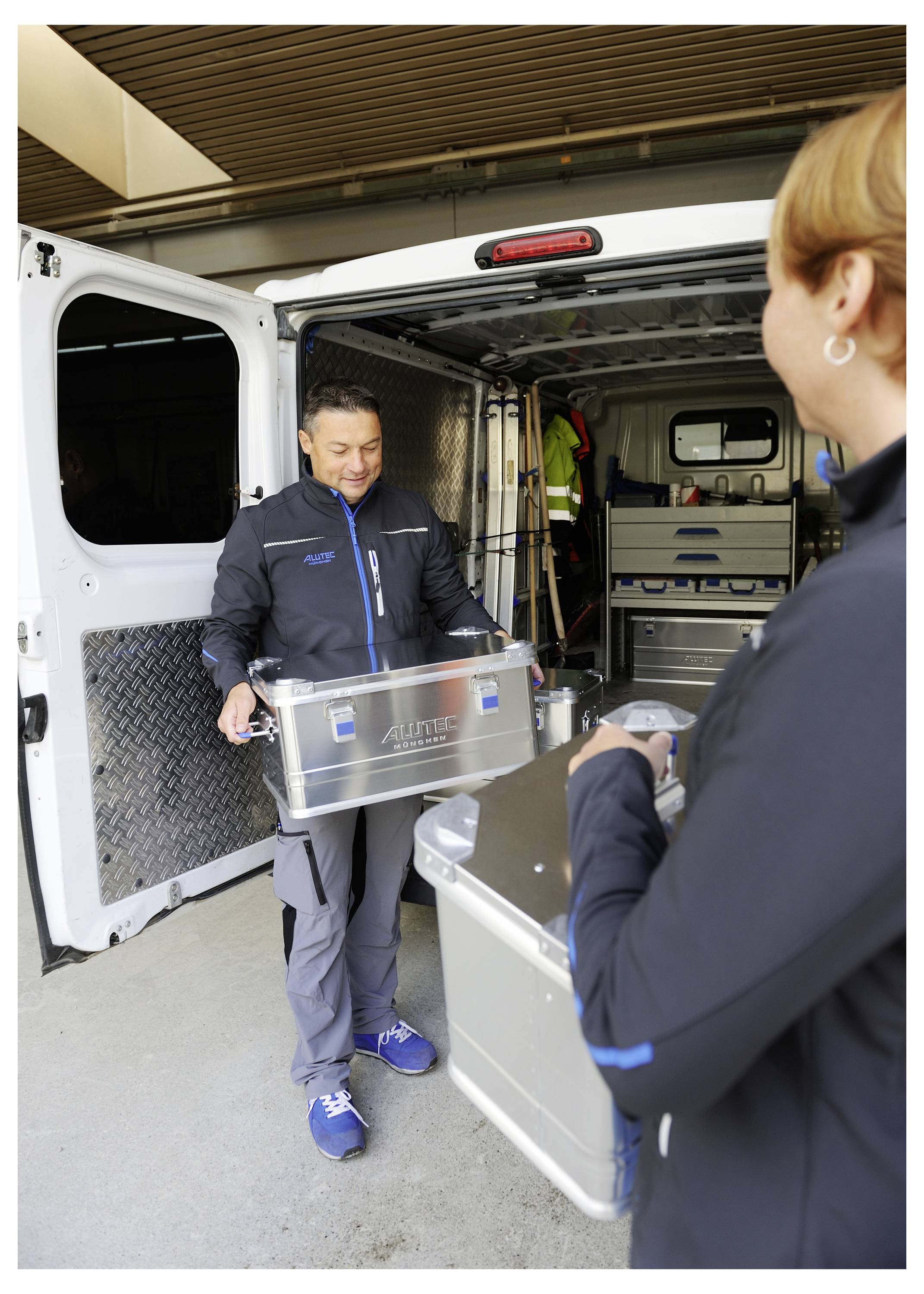 Dwóch pracowników ładuje metalowe walizki do bagażnika furgonu, co sugeruje zadanie transportu lub dostarczania. Drzwi furgonu są otwarte.