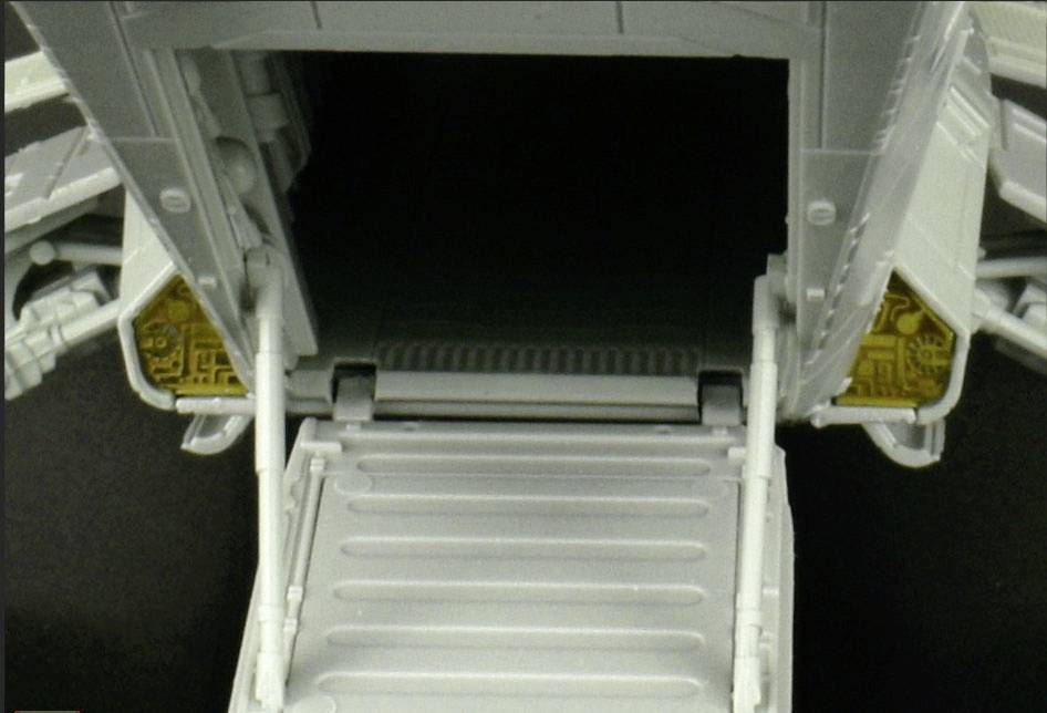 Szara rampa załadowcza, prowadząca do ciemnego pomieszczenia, otoczona technicznymi detalami na obrzeżach.