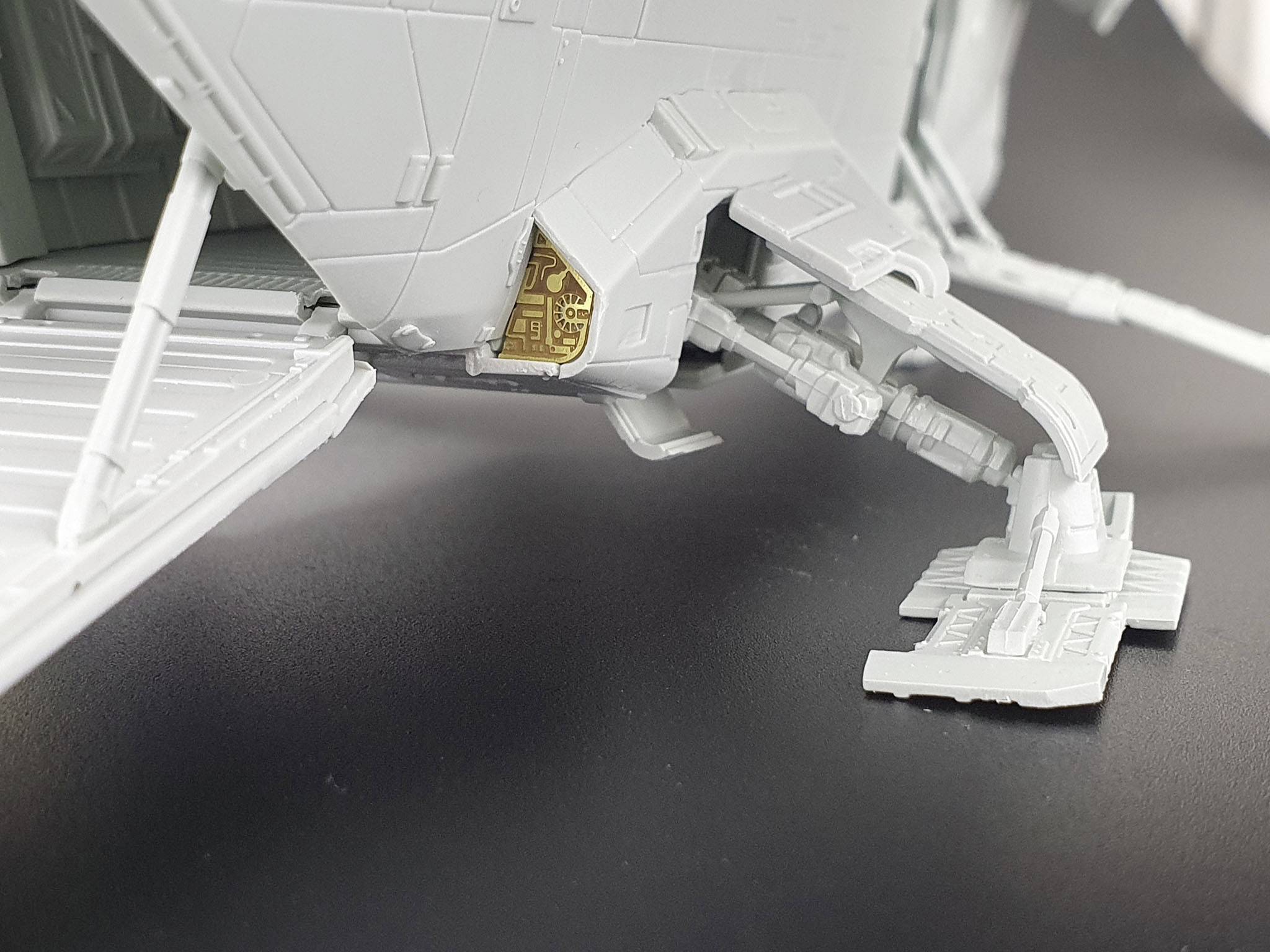 Szary model futurystycznego pojazdu kosmicznego na czarnej powierzchni, ukazujący złożony szczegół podwozia z elementem w kolorze złotym.