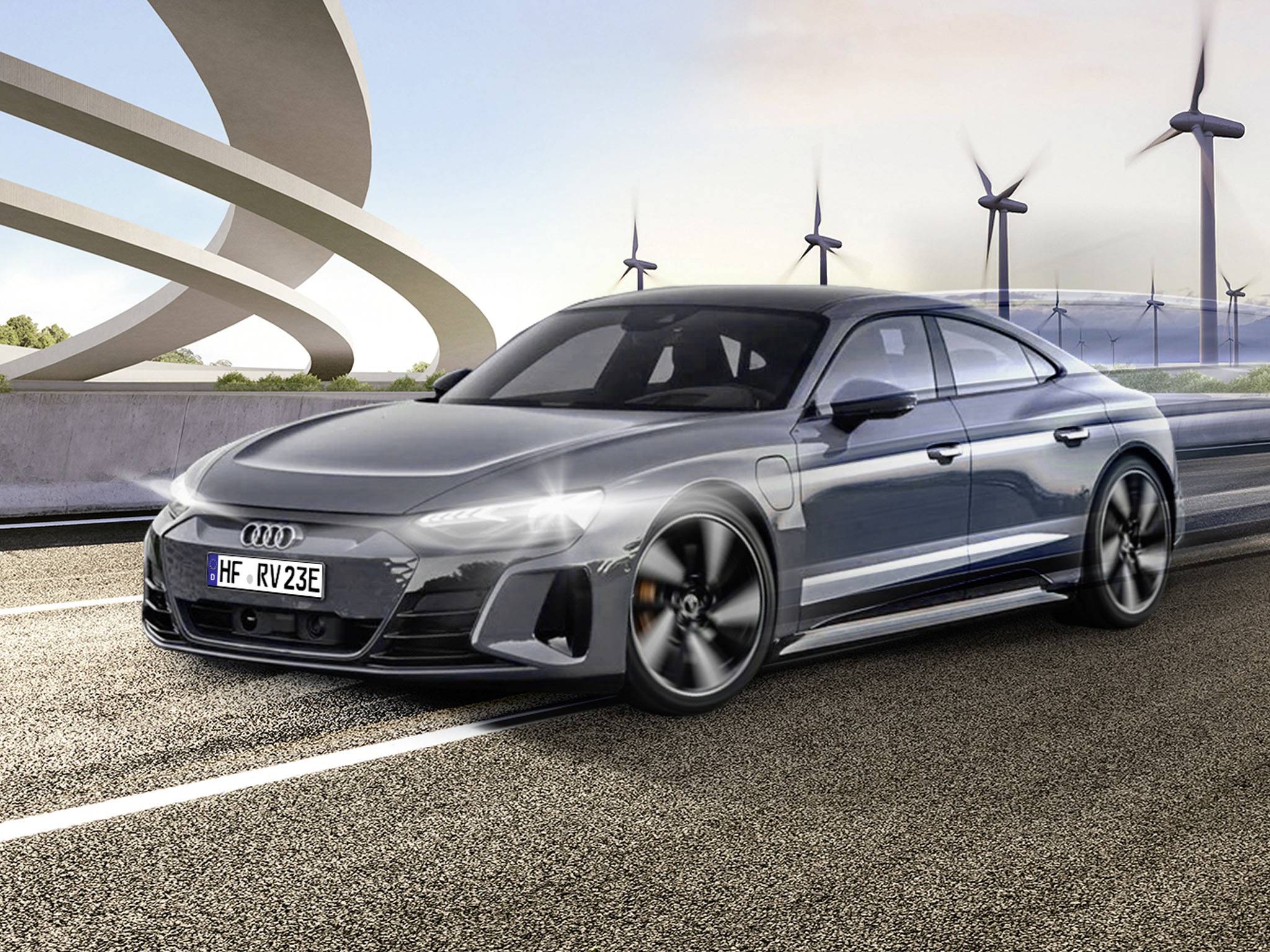 Szary samochód jedzie po drodze, a w tle widać elektrownie wiatrowe, co podkreśla nacisk na ekologiczny transport.