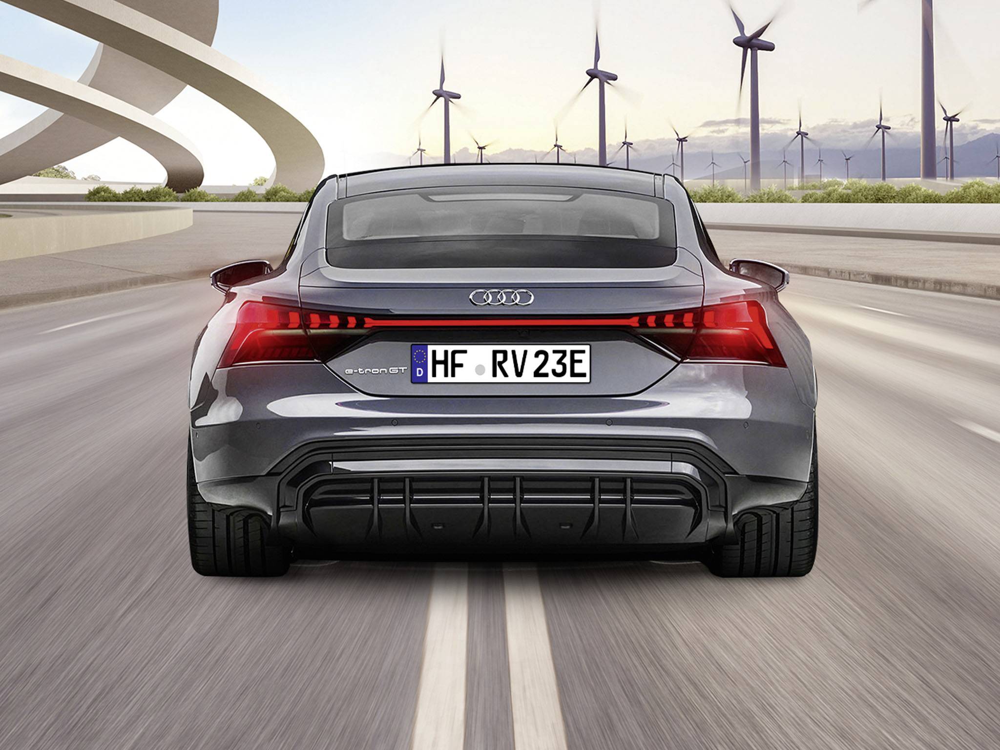 Szary samochód elektryczny jedzie po drodze, otoczony elektrowniami wiatrowymi. Na tablicy rejestracyjnej widnieje napis 'HF - RV23E'.