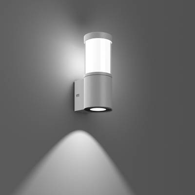 Nowoczesna lampa ścienna z cylindrycznym szklanym kloszem i metalowym mocowaniem rzuca światło w dół na szarą ścianę.
