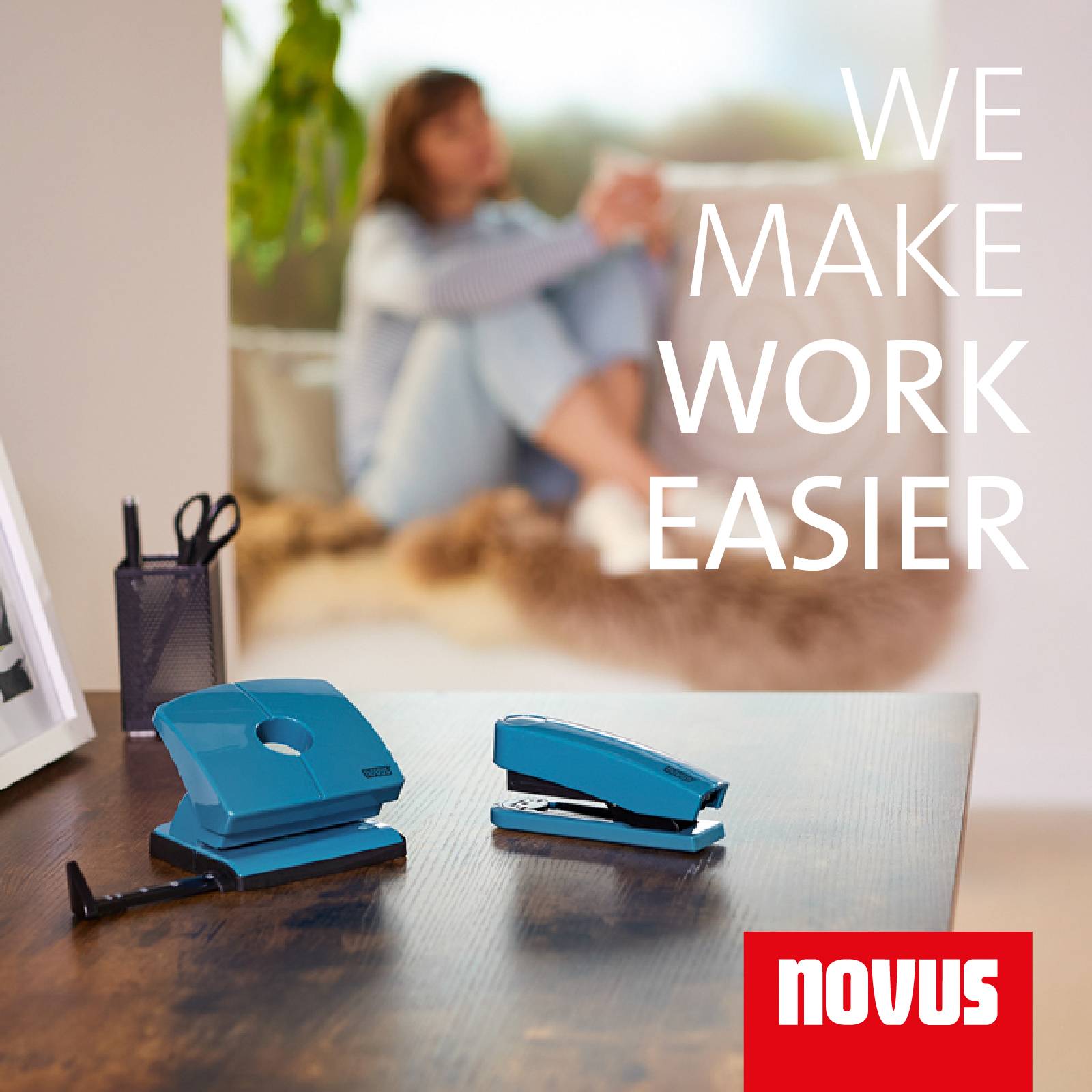 Osoba siedzi spokojnie w tle na kanapie, podczas gdy na pierwszym planie znajduje się niebieski zestaw biurowy ze zszywaczem i dziurkaczem na stole. Tekst na obrazie: 'Ułatwiamy pracę', logo marki: Novus.