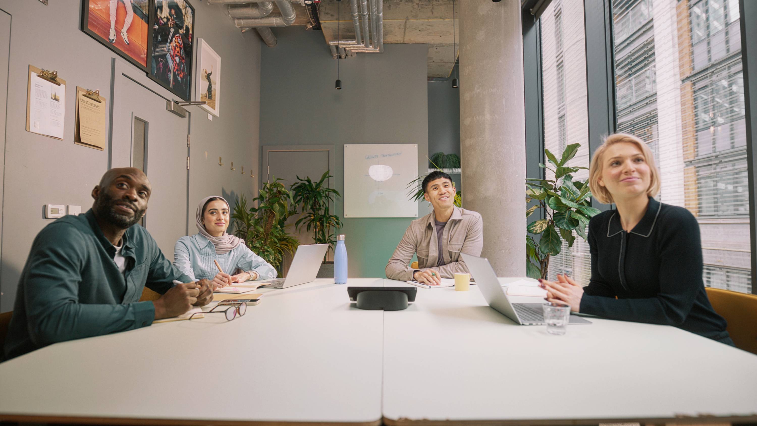 Kilka osób siedzi uśmiechniętych przy stole konferencyjnym z laptopami i notatkami, w nowoczesnym biurze z oknami i roślinami.