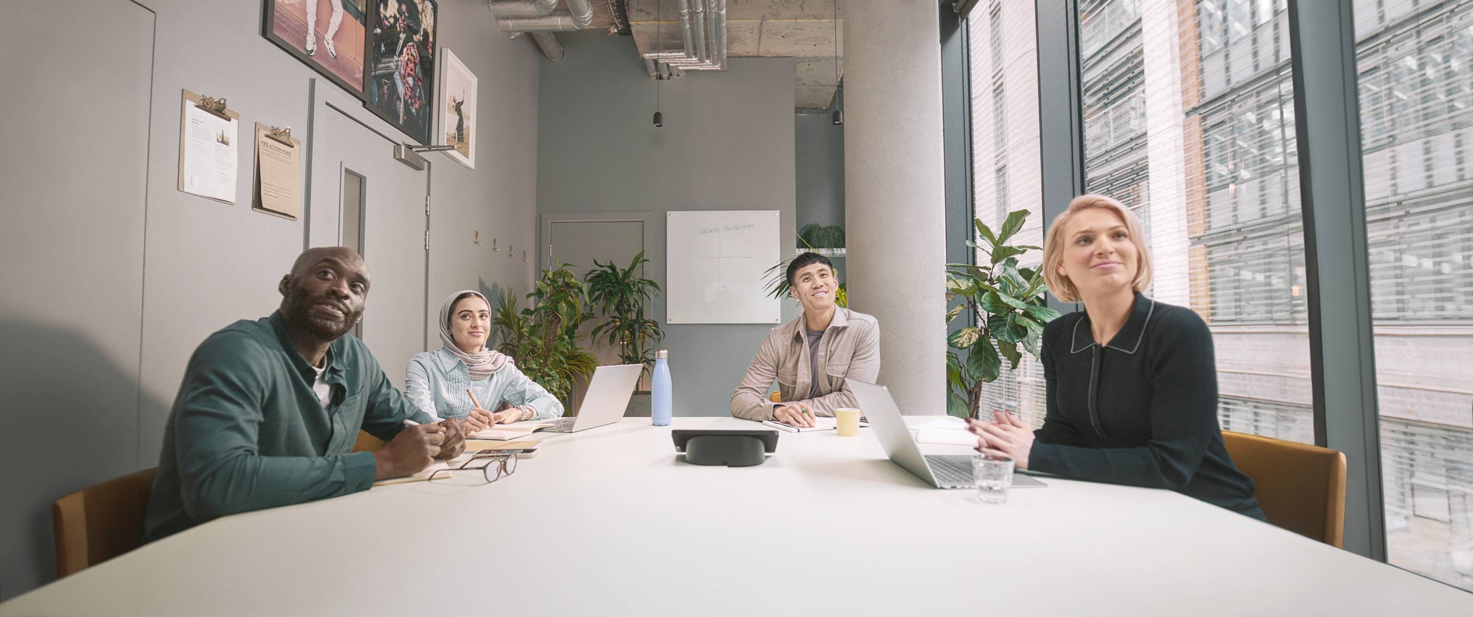 Cztery osoby siedzą przy stole konferencyjnym i zainteresowane patrzą na ekran. W tle widoczne są okna i rośliny.