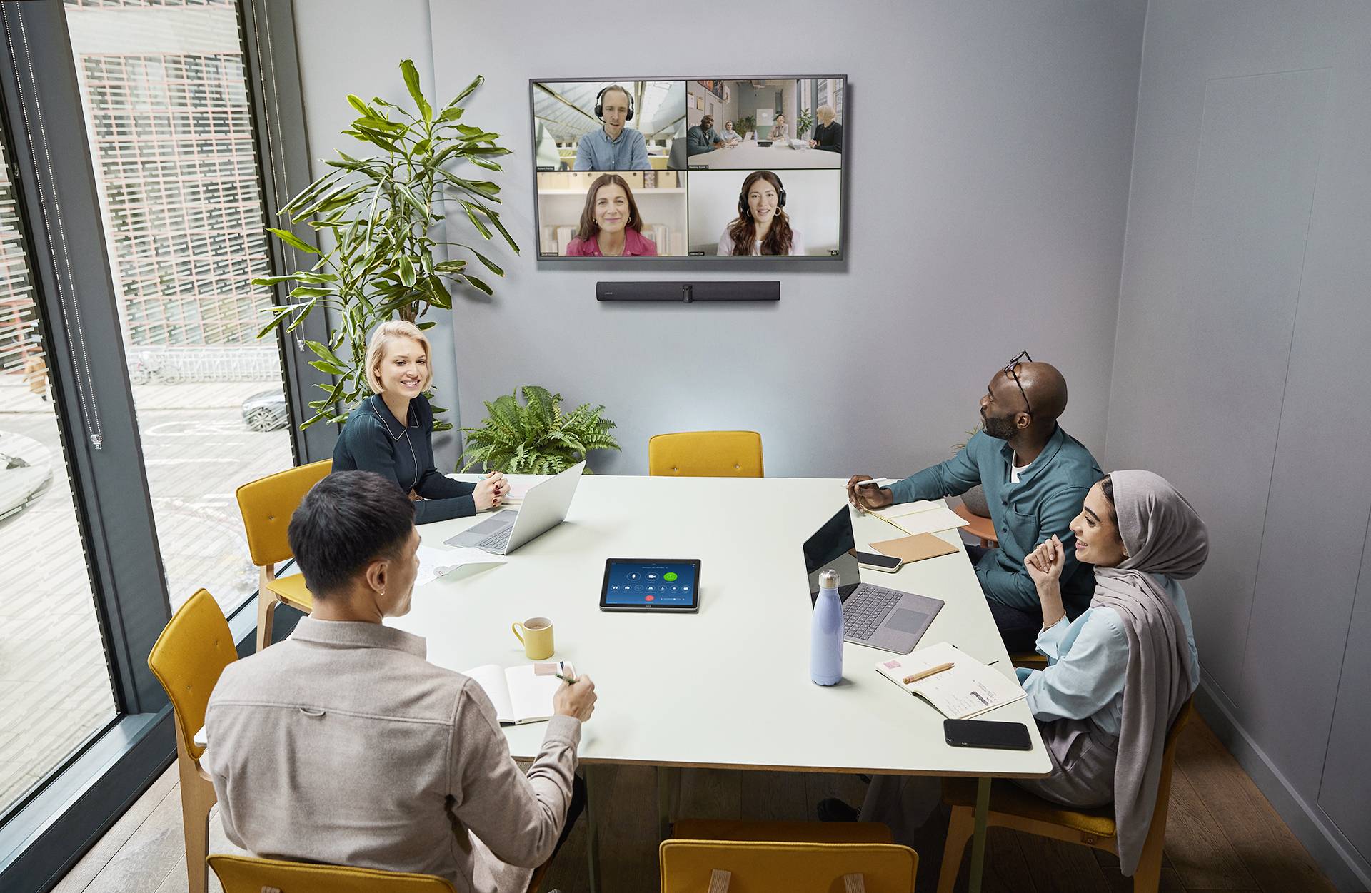 Grupa pięciu osób siedzi przy stole konferencyjnym i bierze udział w wideokonferencji z czterema osobami wyświetlanymi na ekranie.