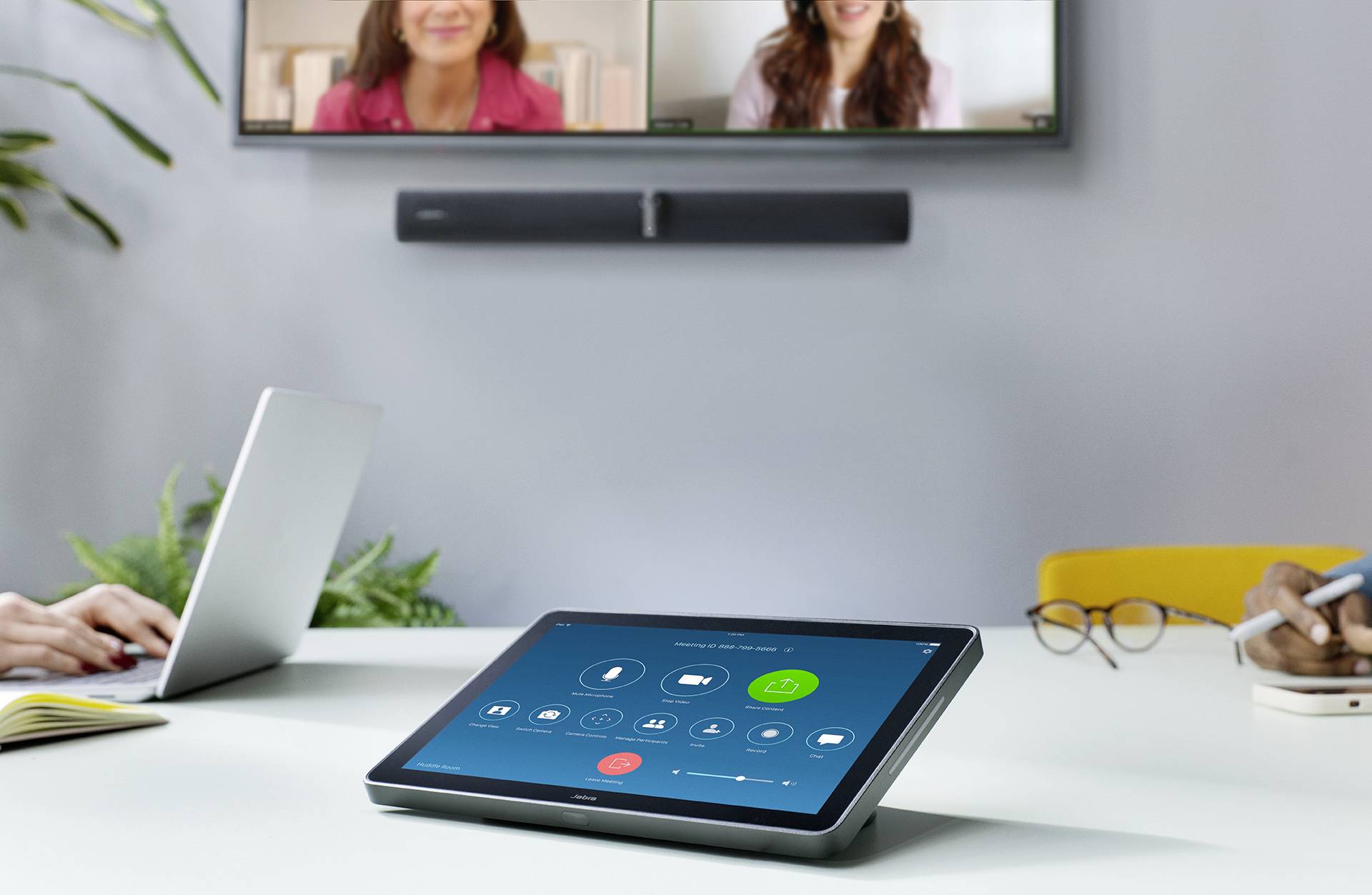 Stół konferencyjny z tabletem na pierwszym planie, sterującym wideorozmową. W tle ekran z dwiema osobami podczas wideokonferencji.