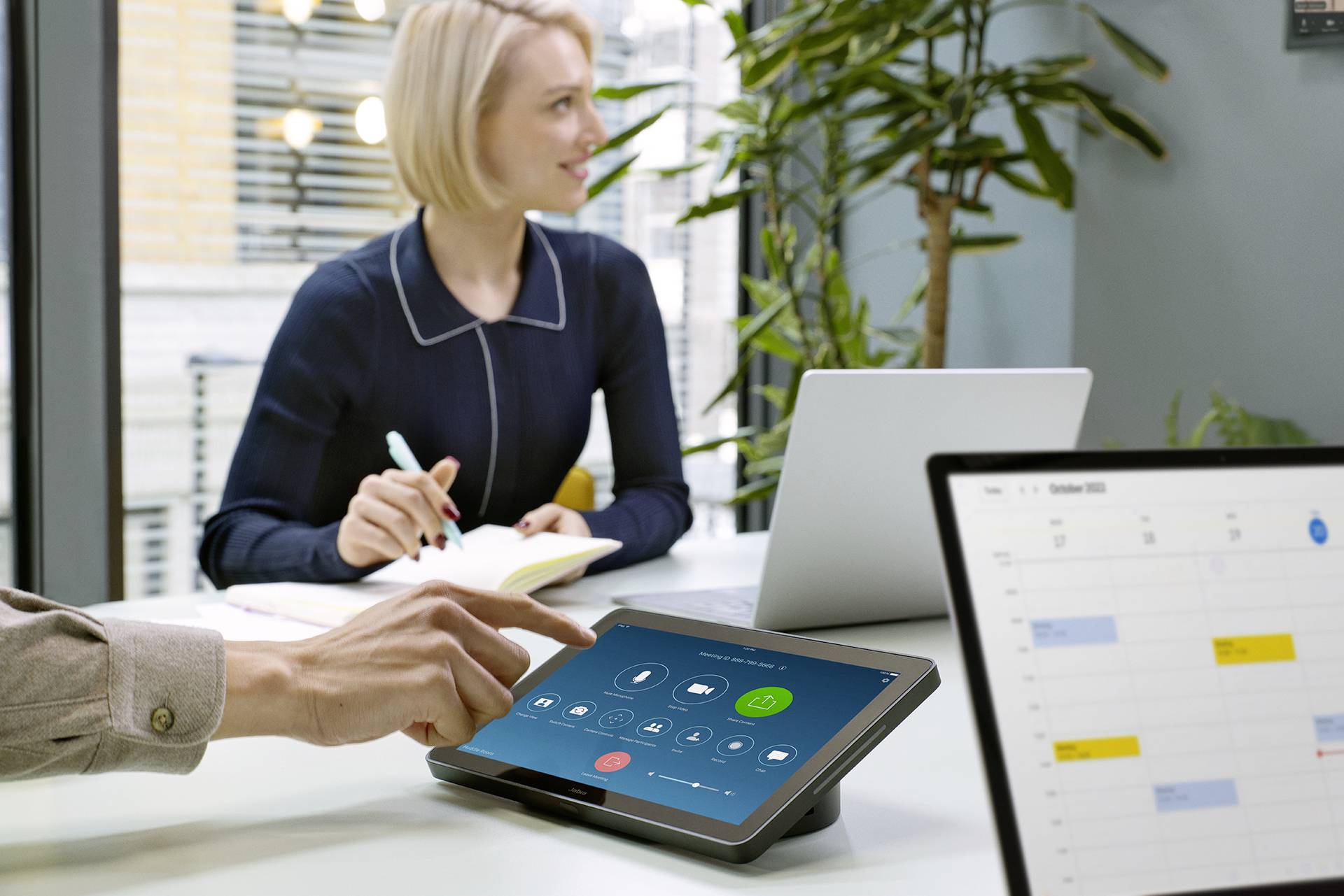 Osoba obsługuje tablet z oprogramowaniem do wideokonferencji, podczas gdy w tle kobieta sporządza notatki. Na laptopie widoczny jest kalendarz.