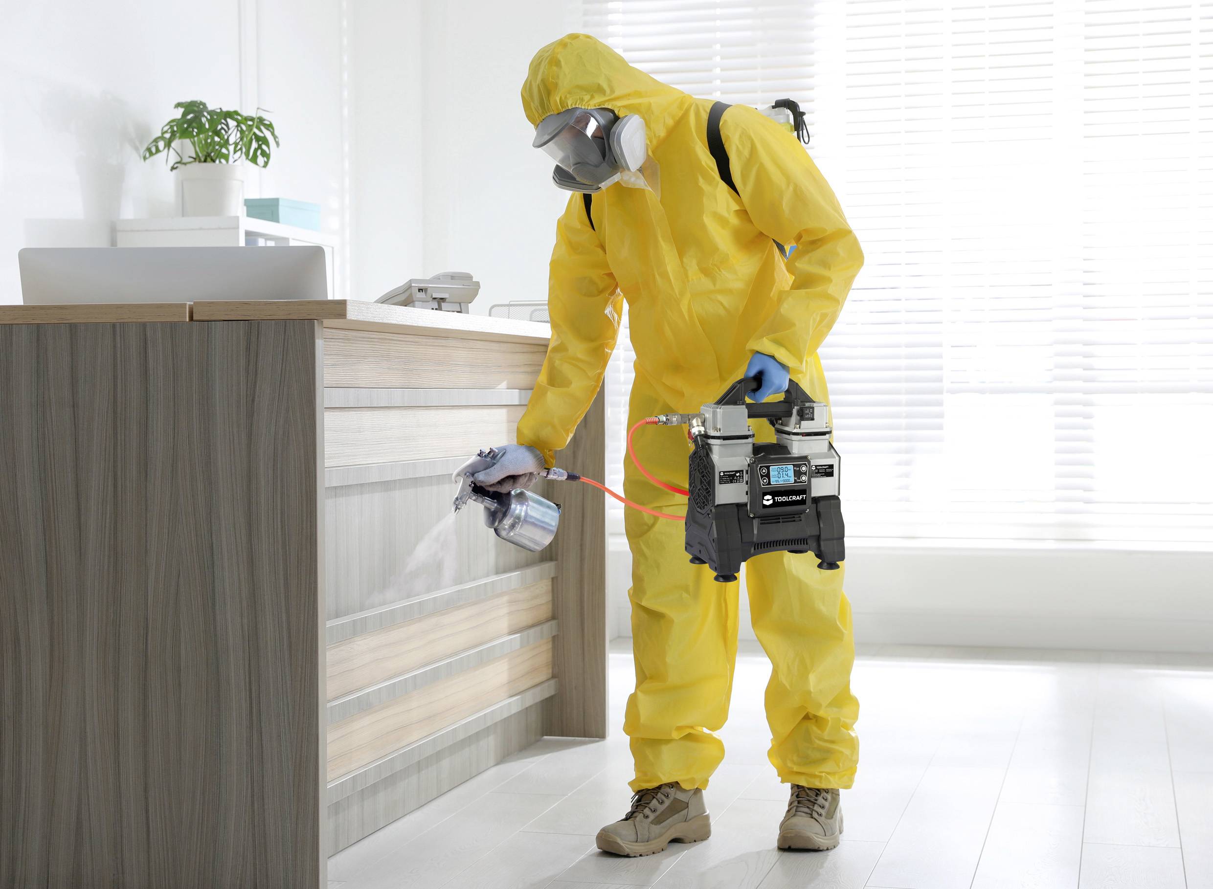 Osoba w żółtym kombinezonie ochronnym dezynfekuje powierzchnię za pomocą urządzenia natryskowego w biurze. W tle jasne okna.