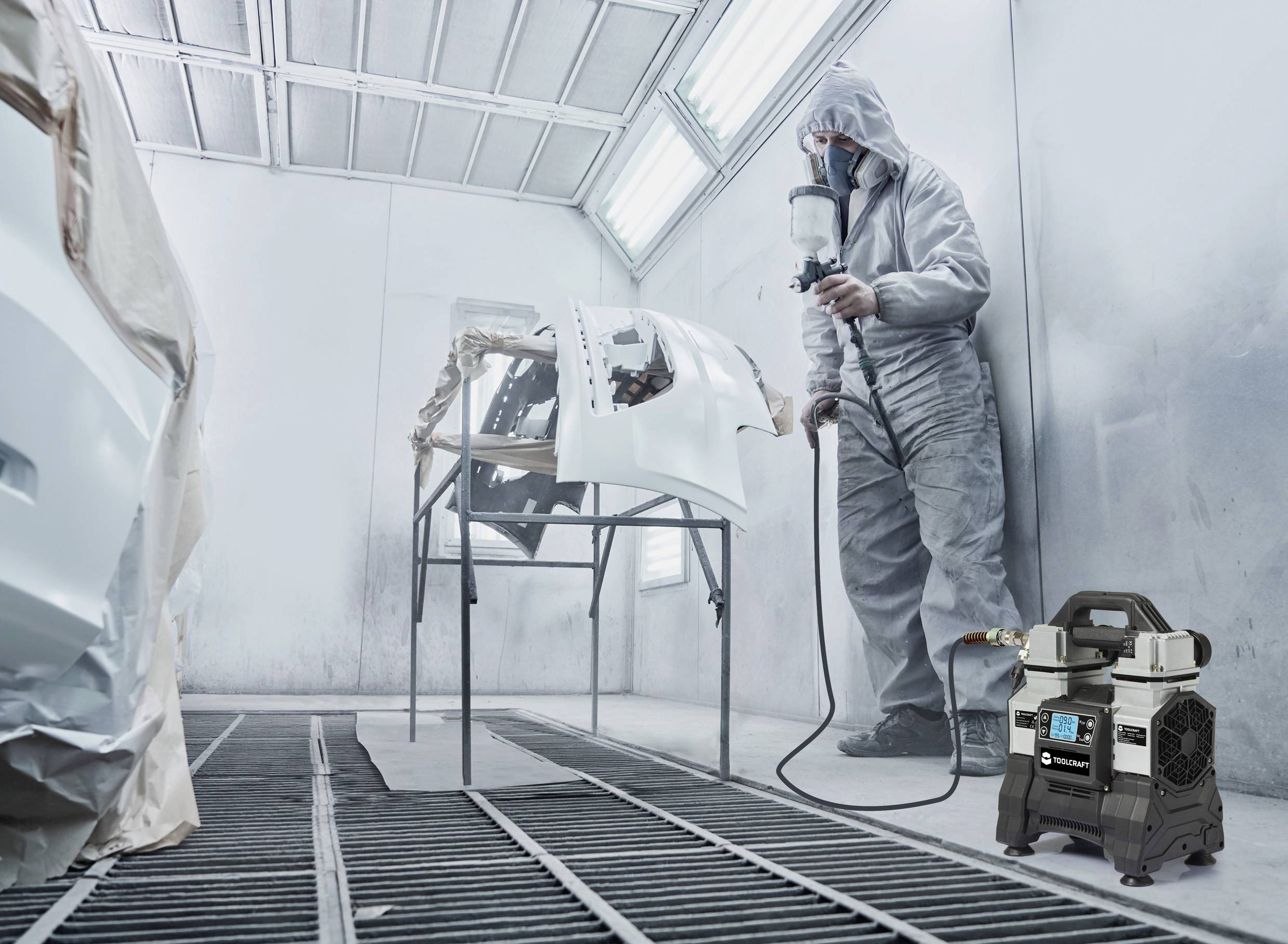 Pracownik w kombinezonie ochronnym maluje części samochodowe w kabinie lakierniczej. W tle widoczny jest pistolet natryskowy i sprężarka powietrza.