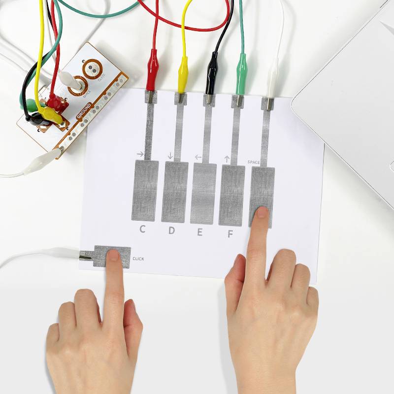 Dwie ręce na kartce z klawiszami strzałek i literami (C, D, E, F) połączonymi krokodylkami z płytką Makey Makey.