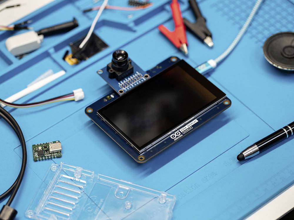 Urządzenie Arduino z wyświetlaczem i kamerą leży na niebieskiej macie roboczej, otoczone akcesoriami elektronicznymi i narzędziami.