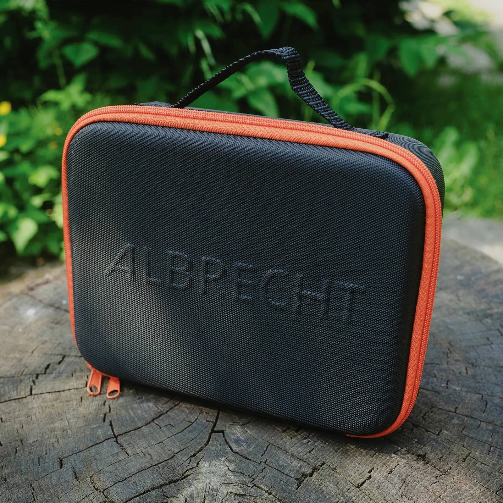 Czarna osłona ochronna z czerwonym zamkiem błyskawicznym i uchwytem do przenoszenia, umieszczona na pniu drzewa. Nazwa 'ALBPECHT' jest wytłoczona na przedniej stronie.