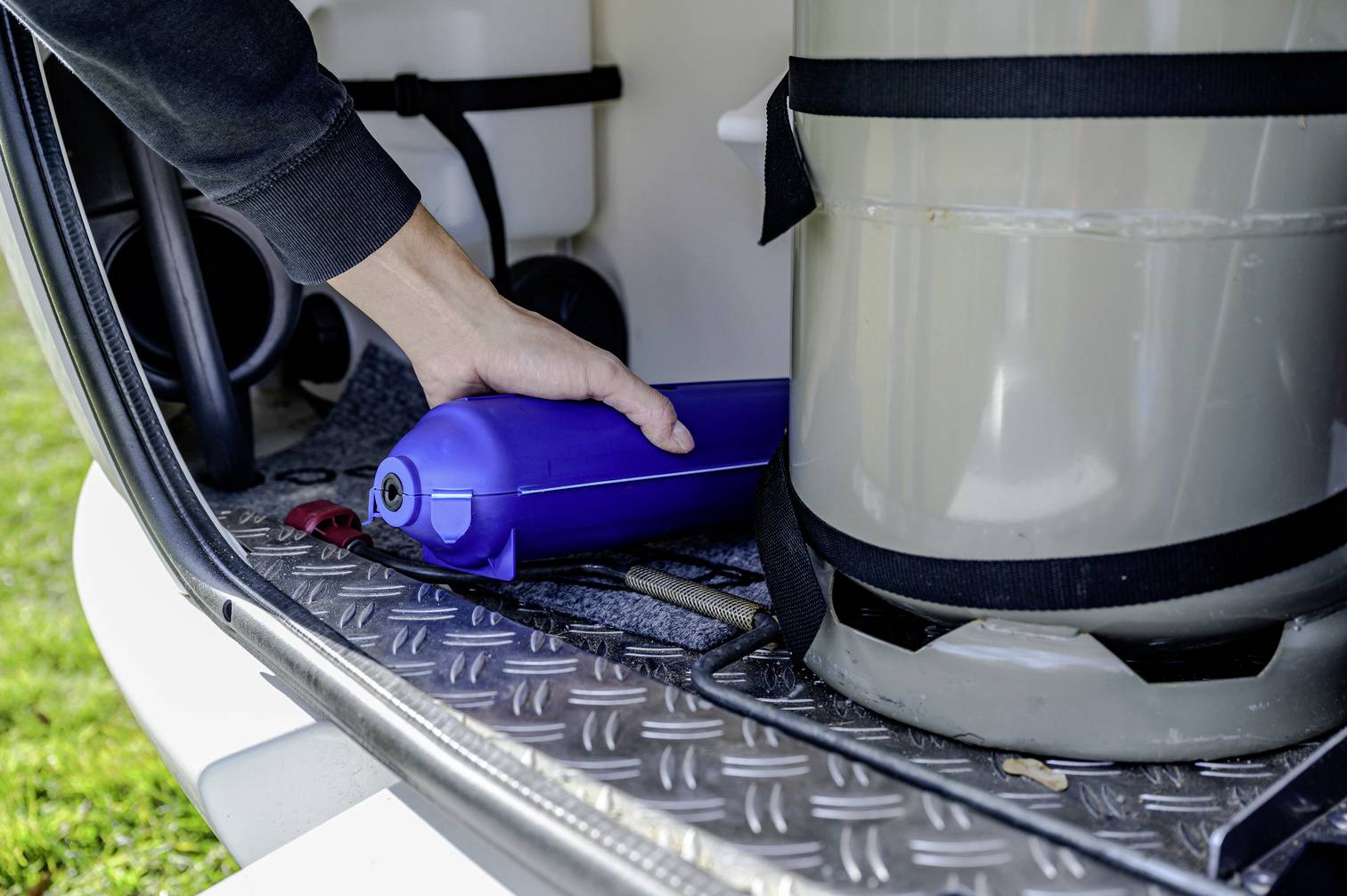 Osoba wkłada niebieski skrzynkę narzędziową do bagażnika obok srebrnego kanistra. W tle widać trawę.
