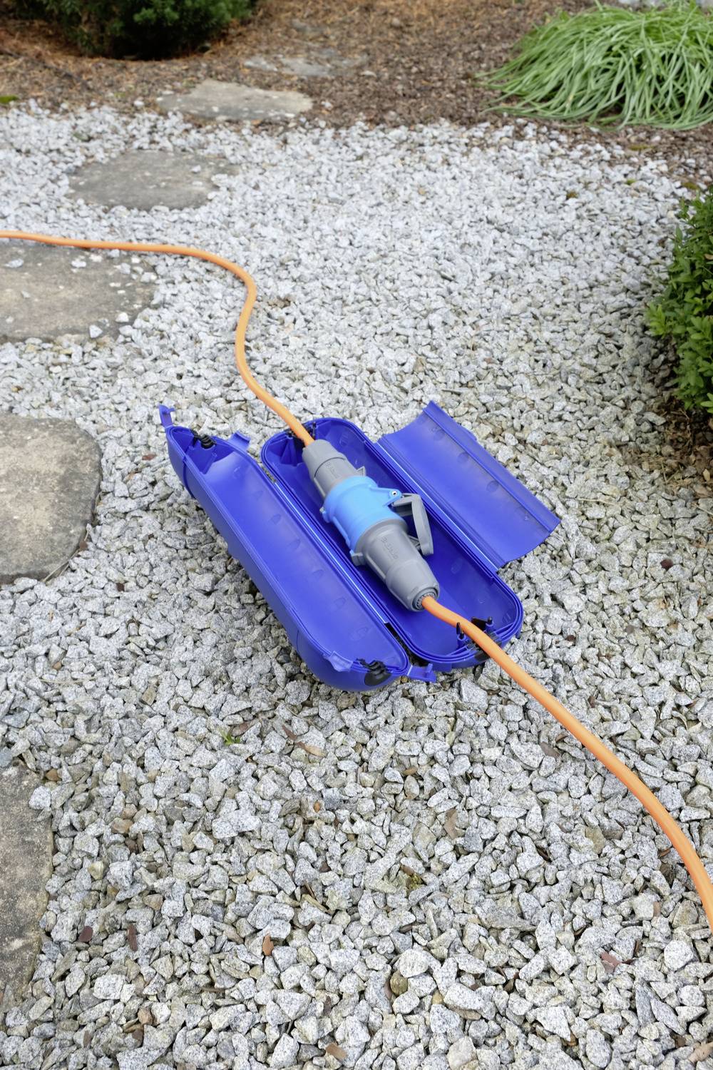 Niebieski osłona kabla leży na żwirowej ścieżce. Pomarańczowy kabel przechodzi przez osłonę. Otoczenie: ogród z kamieniami i roślinami.