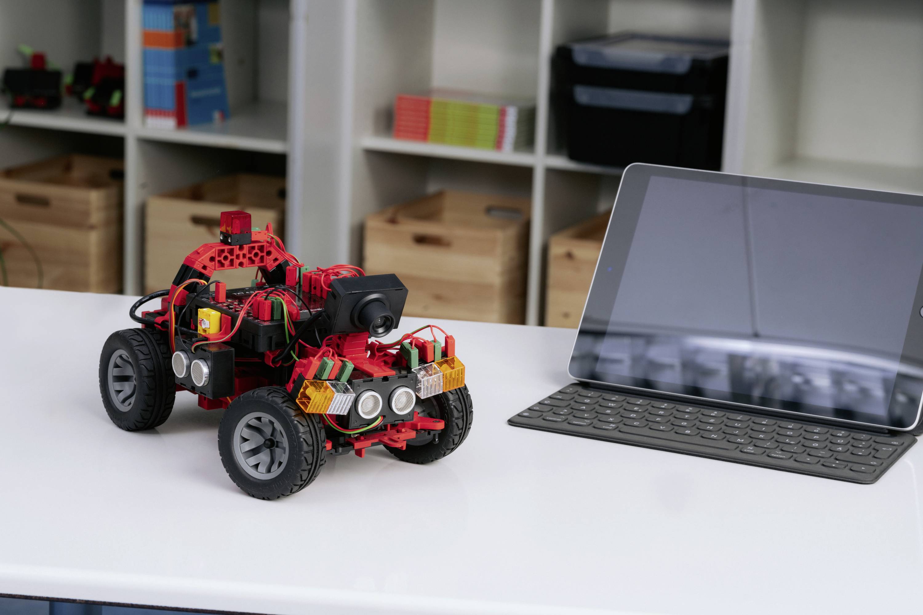 Małe zdalnie sterowane pojazd robotyczny wykonany z tworzywa sztucznego z systemem kamerowym stoi na stole obok tabletu z klawiaturą. W tle widoczne są regały.