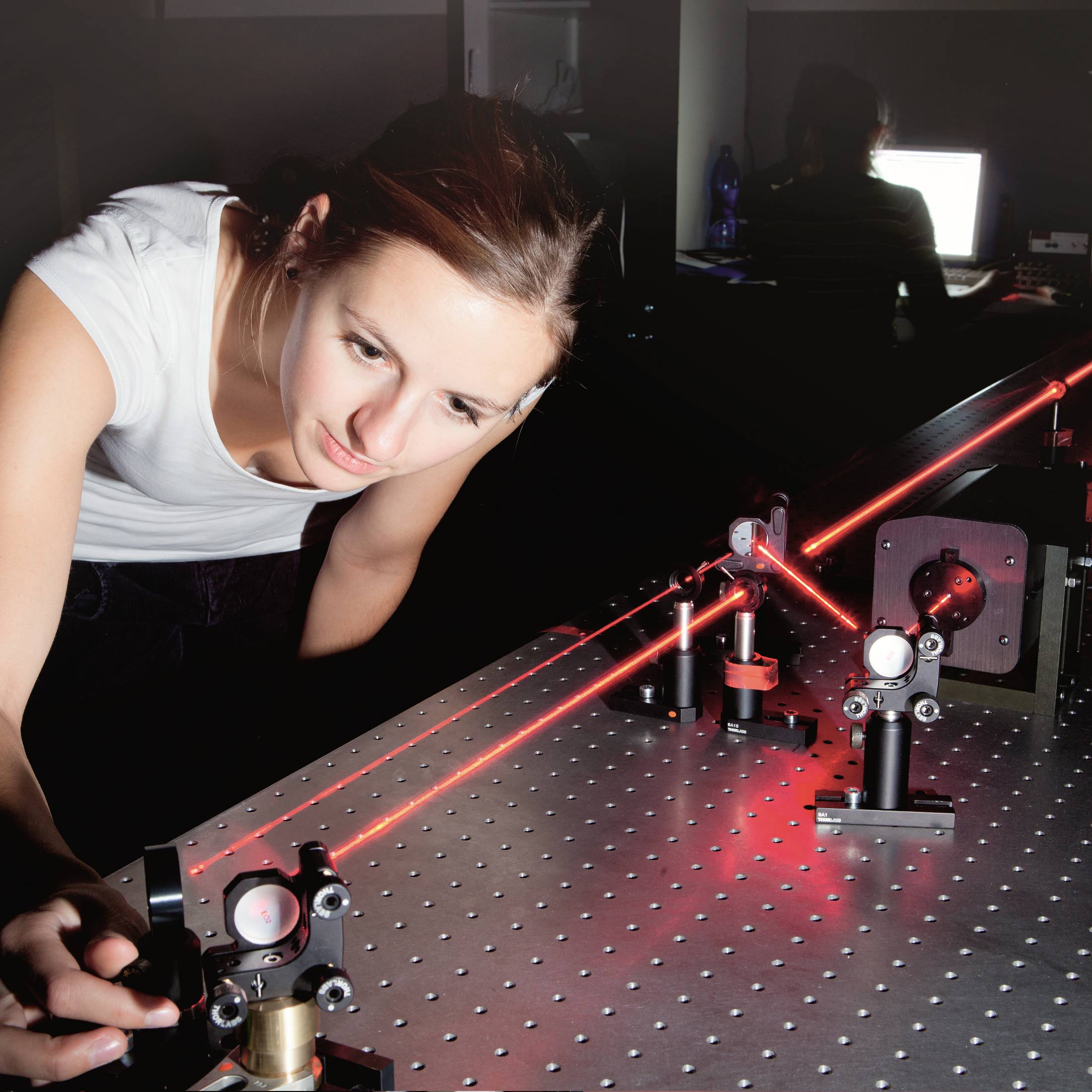 Kobieta pracuje w laboratorium, gdzie laser jest kierowany przez kilka soczewek. W tle świeci ekran komputera.