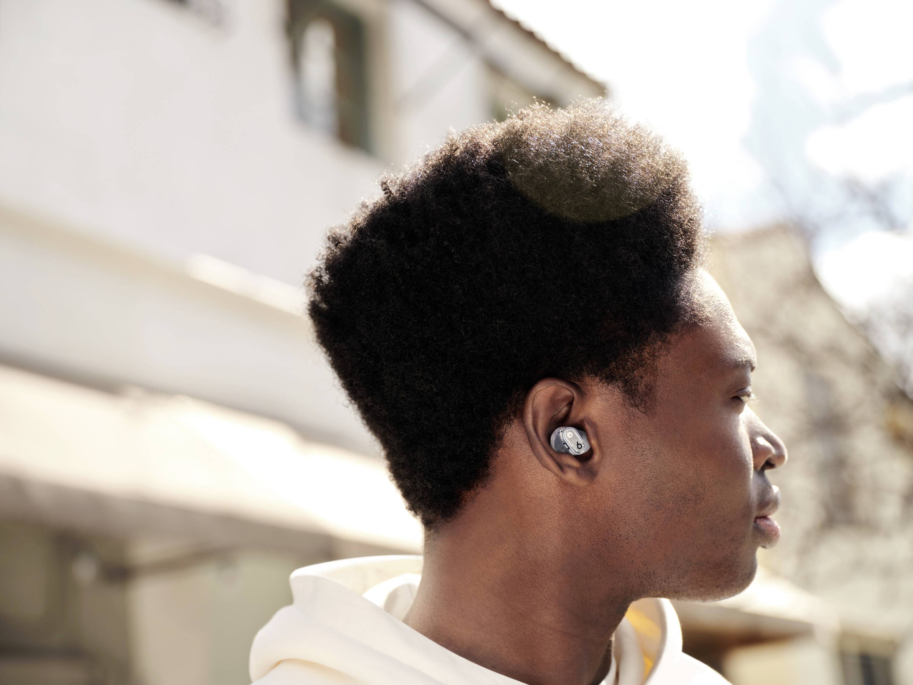 Osoba nosząca słuchawki patrzy w prawo, stojąc na słonecznym terenie zewnętrznym, ze szczególnym skupieniem na słuchawkach.