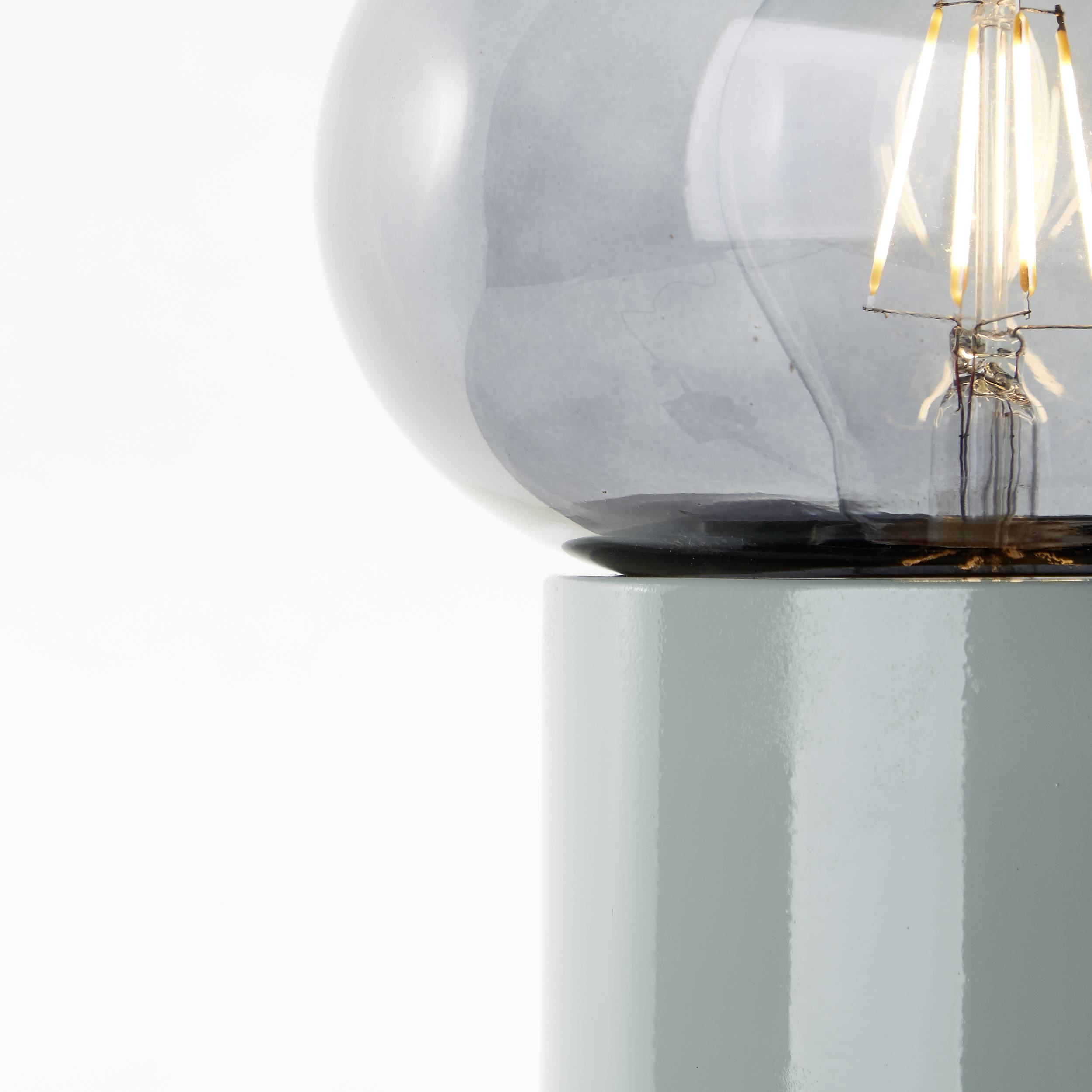 Jasnosiwy, okrągły klosz szklany spoczywa na szarej podstawie metalowej. Żarówka wewnątrz jest widoczna.