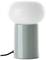 Biała lampa stołowa z okrągłym, błyszczącym kloszem i cylindryczną, zieloną podstawą stoi na podłożu. Czarny kabel jest widoczny.
