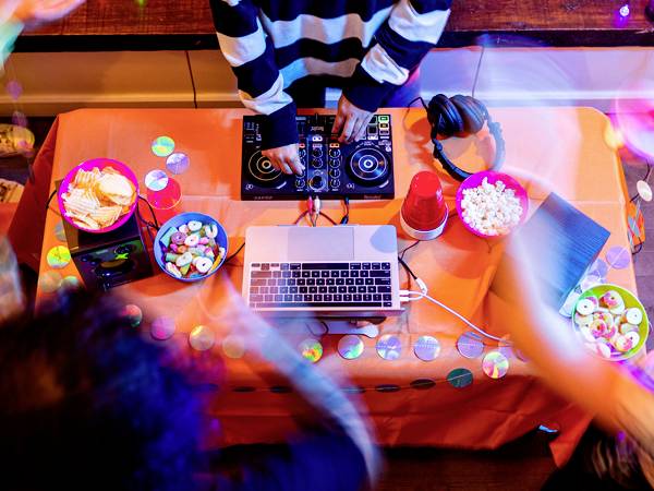 Widok z góry na stół z osprzętem DJ-skim, przekąskami i laptopem. Jedna osoba obsługuje konsolę DJ-ską, podczas gdy na stole rozłożone są przekąski.