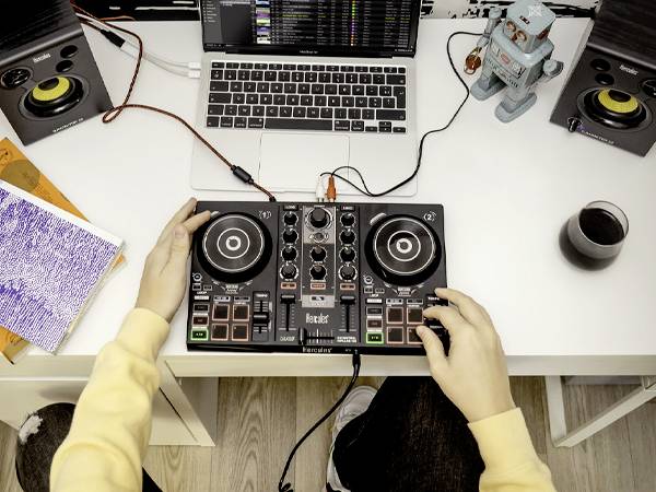 'Osoba miesza muzykę na kontrolerze DJ-skim na biurku z laptopem, głośnikami, notatnikiem, robotem zabawkowym i napojem.'