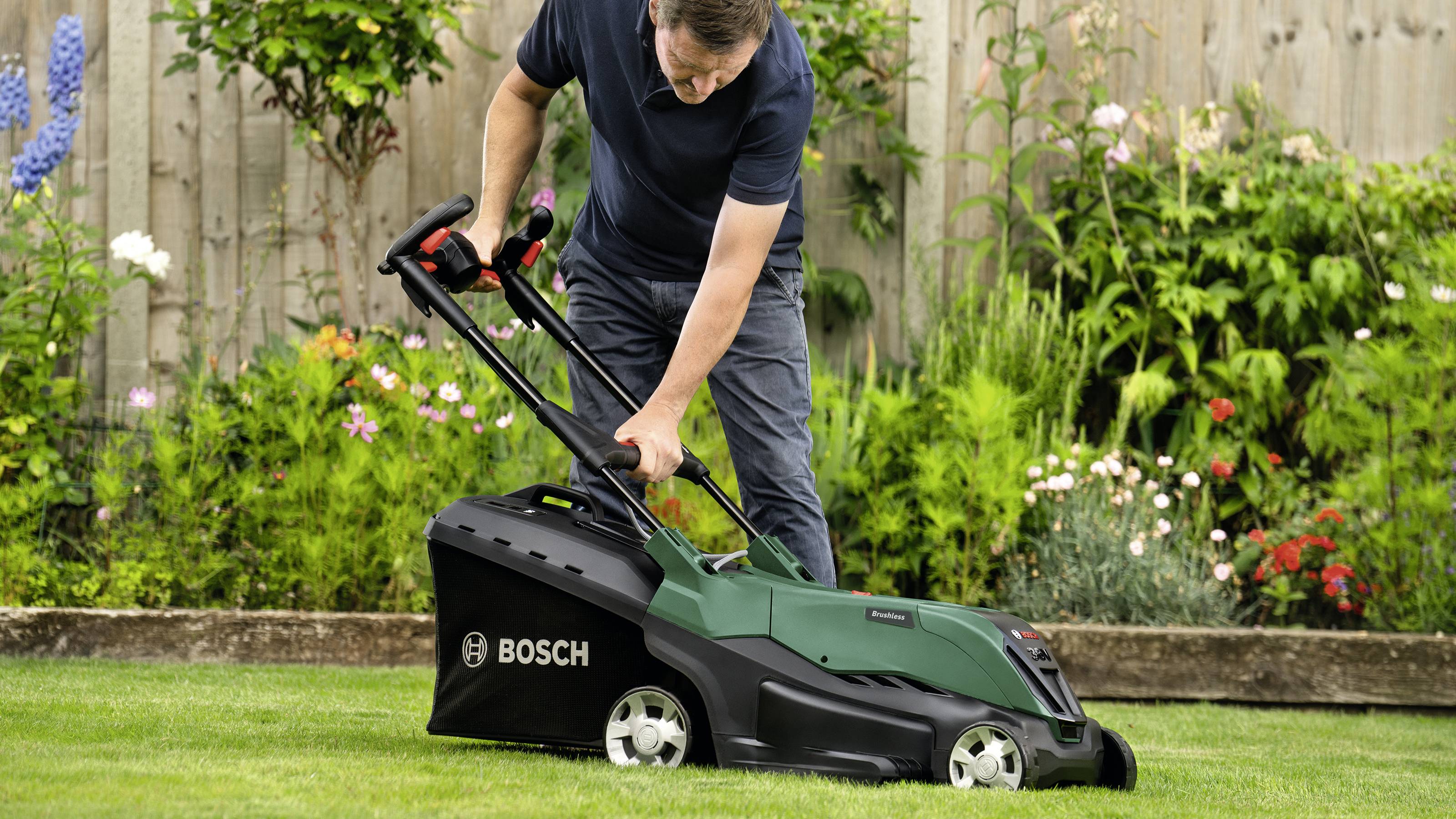 Osoba kosi trawę zielonym kosiarką Bosch w ogrodzie. W tle widoczne są rabaty kwiatowe i drewniany płot.