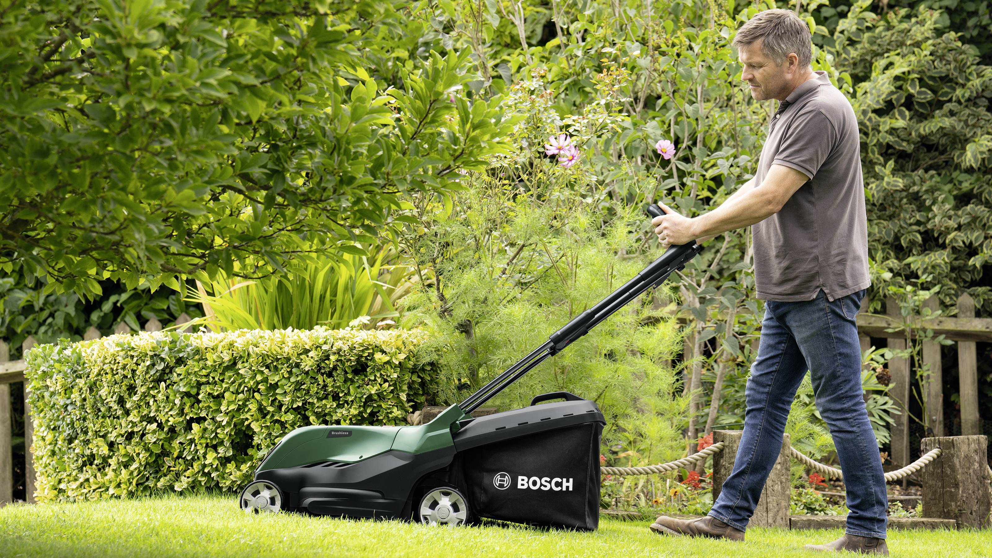Mężczyzna kosi trawę zielonym kosiarką Bosch w zielonym ogrodzie pełnym roślin i krzewów.