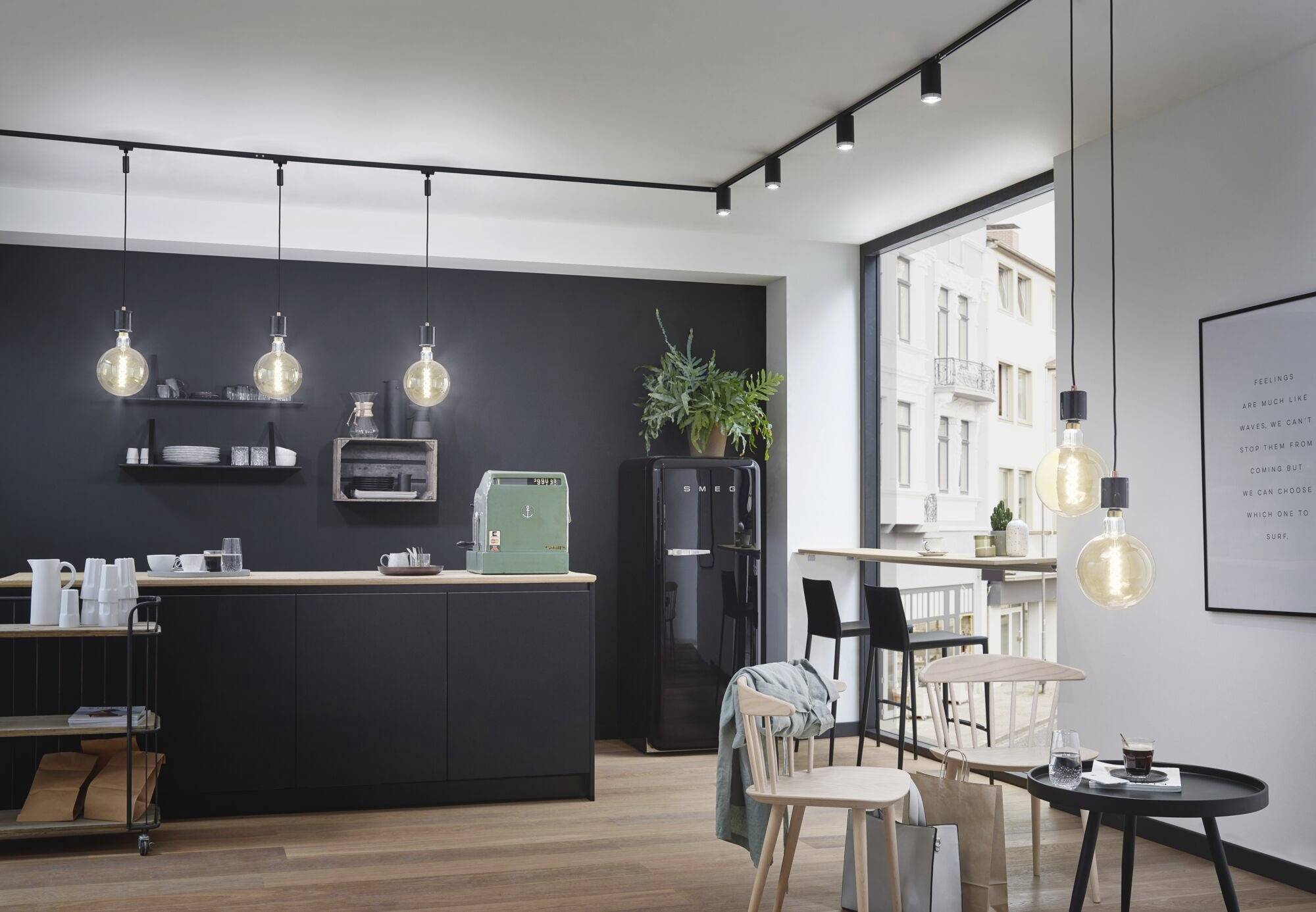 Nowoczesna kuchnia z czarną wyspą kuchenną i wiszącymi żarówkami. W tle widoczny jest lodówka, okna i rośliny.