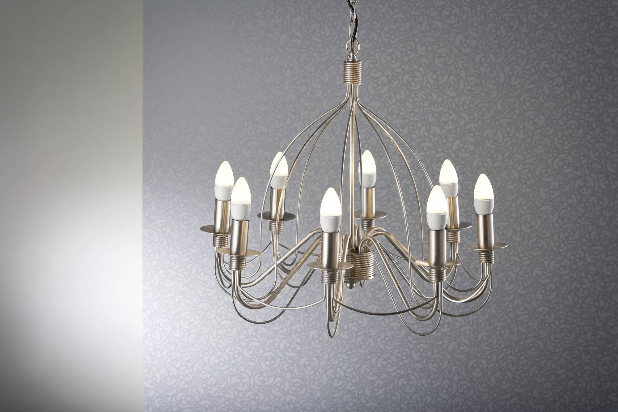 Elegancki żyrandol z dziewięcioma świecznikowymi lampami wisi od sufitu na tle dyskretnej, wzorzystej tapety.