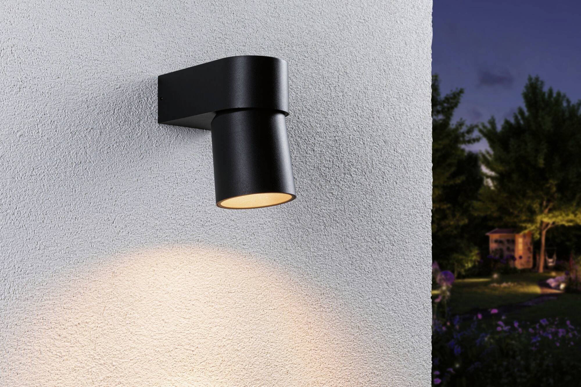 Lampa na ścianie zewnętrznej z nowoczesnym czarnym mocowaniem, delikatnie oświetlająca strukturyzowaną białą ścianę; nastrój wieczoru w tle.