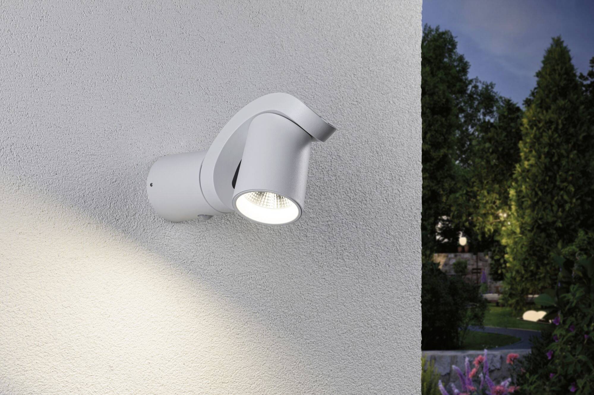 Biała lampa ścienna zamontowana na ścianie budynku, oświetlająca obszar poniżej. W tle widoczny ogród w wieczornym świetle.