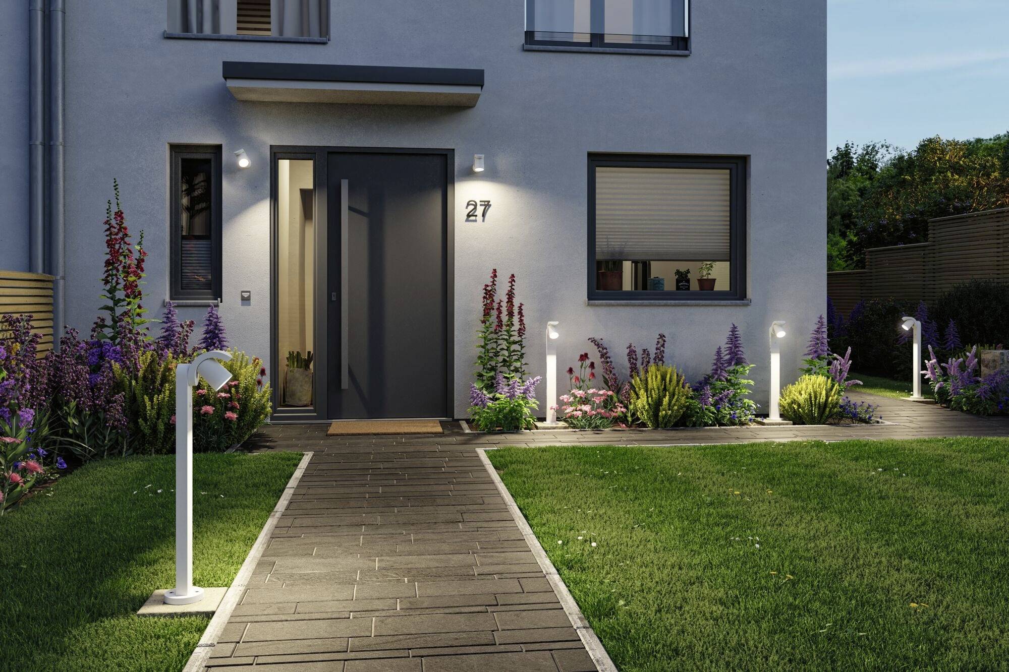 Nowoczesny dom nocą, oświetlone wejście, numer domu '27'. Rabatki kwiatowe po bokach ścieżki, delikatne oświetlenie ogrodu.