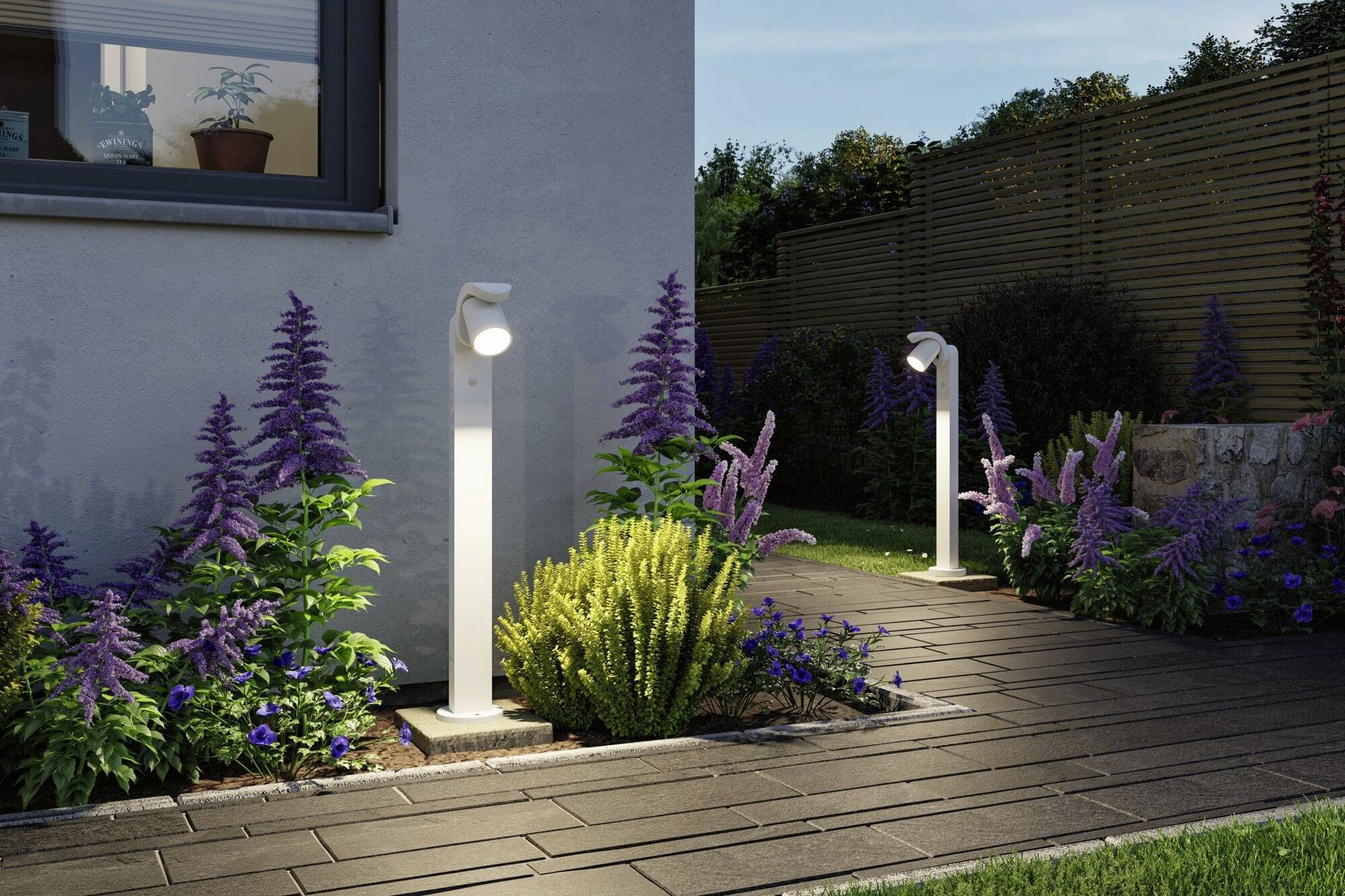 Ścieżka ogrodowa z nowoczesnym oświetleniem, otoczona kwiatami w odcieniach fioletu i zieleni, biegnąca obok domu o zmierzchu.