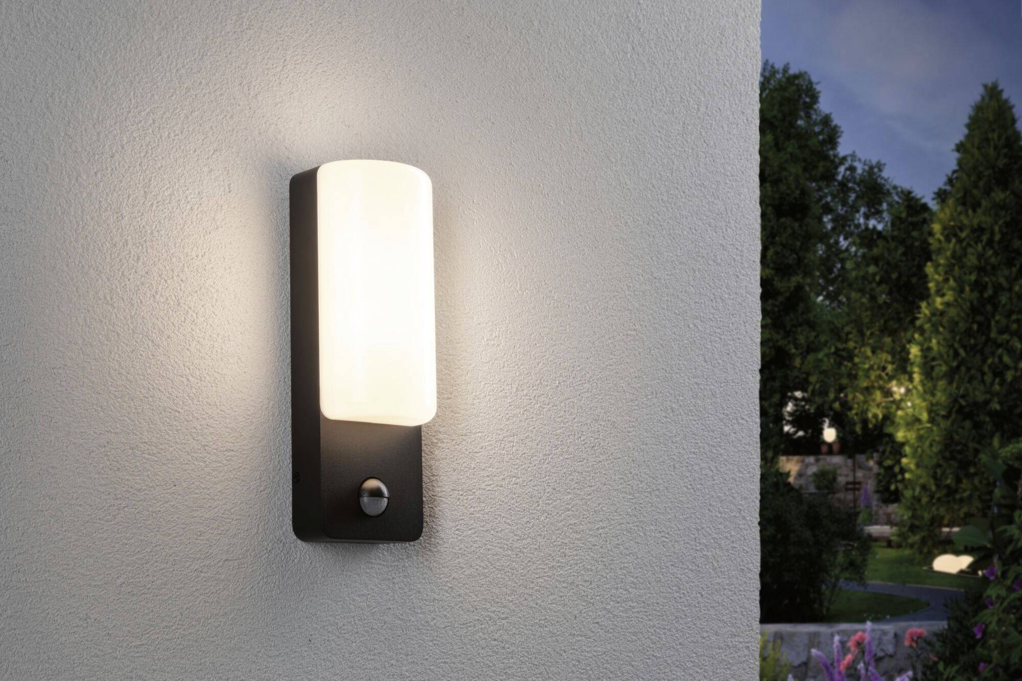 Lampa zewnętrzna na ścianie o zmierzchu, dająca ciepłe światło i oświetlająca obszar wokół domu, otoczona drzewami w tle.