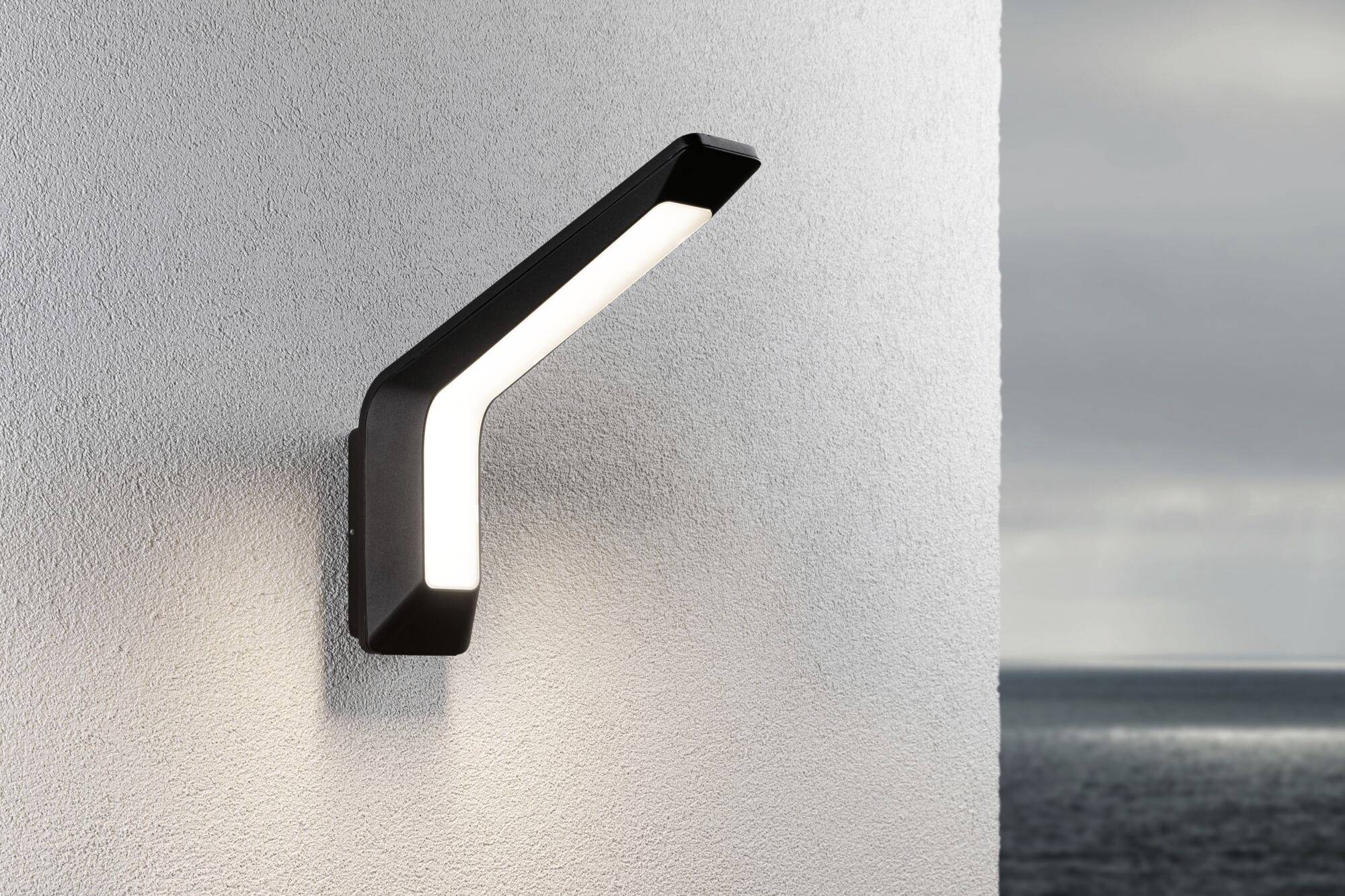 Nowoczesna lampa ścienna w kształcie litery Z zamontowana na zewnętrznej ścianie, wieczorem oświetla strukturalną białą ścianę. W tle widok na morze.