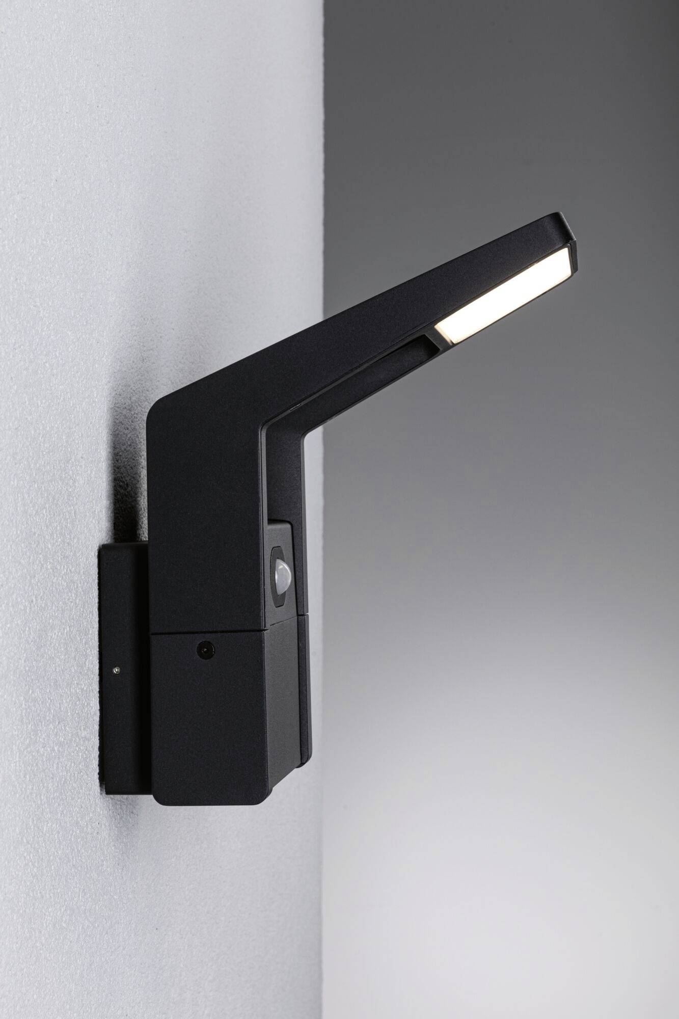 Nowoczesna lampa zewnętrzna z czujnikiem montowana na ścianie. Czarny, minimalistyczny design z prostokątną głowicą świetlną. Oświetla fragment ściany.