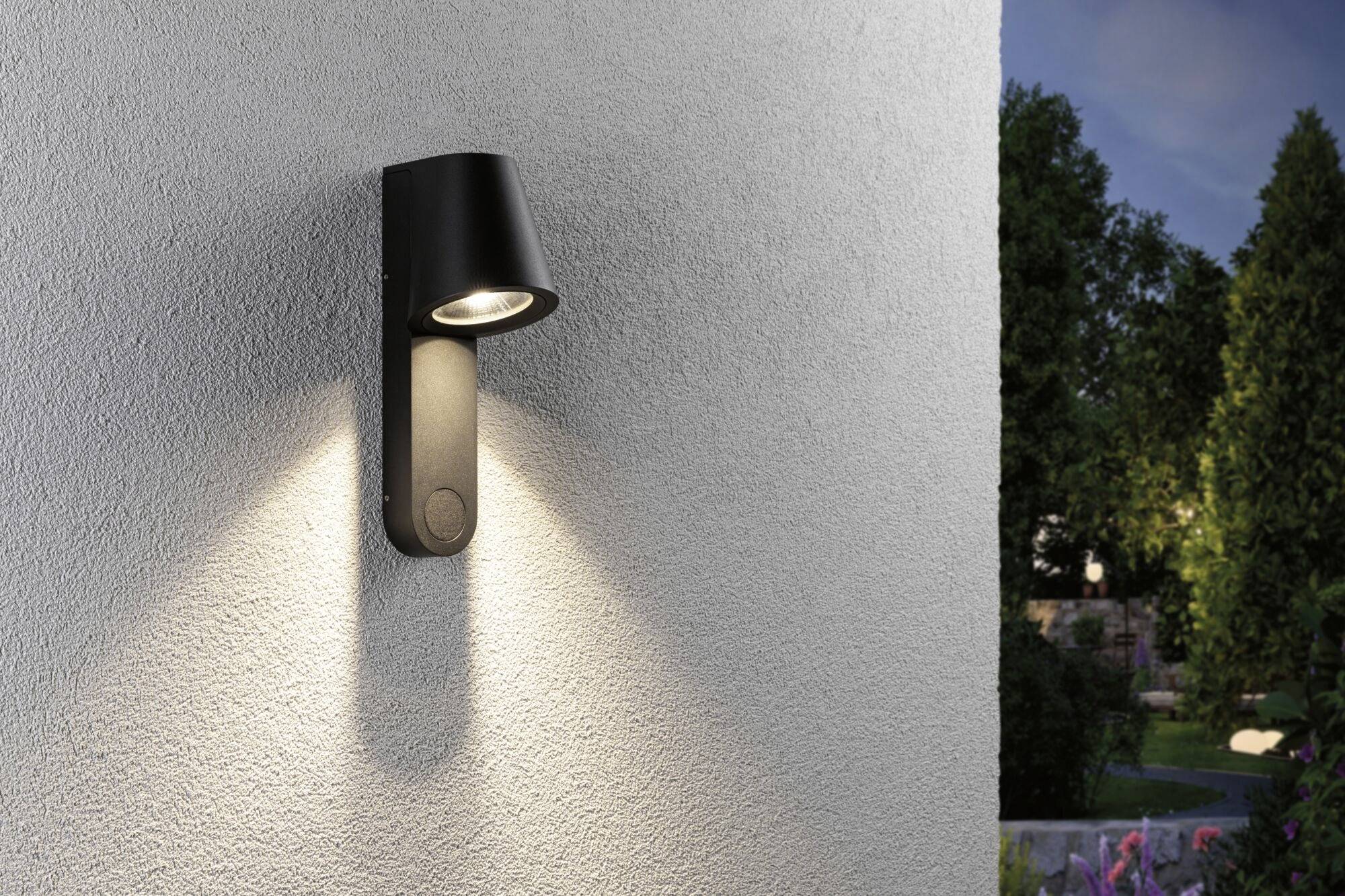 Kinkiet na zewnętrznej ścianie na szarym murze, oświetlający krajobraz ogrodu o zmierzchu.