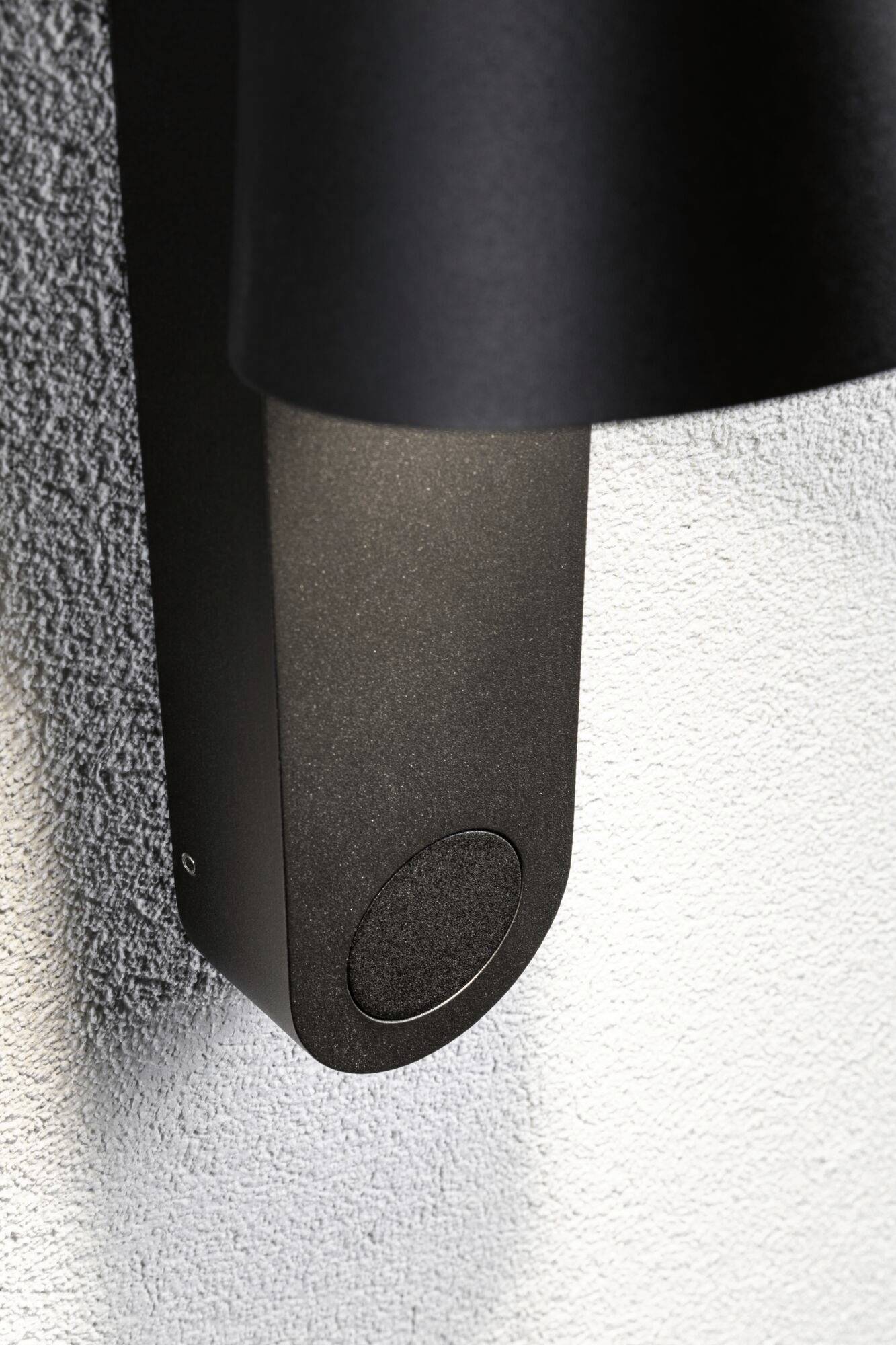 Czarna lampa zewnętrzna na szarej ścianie, prostokątna z zaokrąglonymi narożnikami.