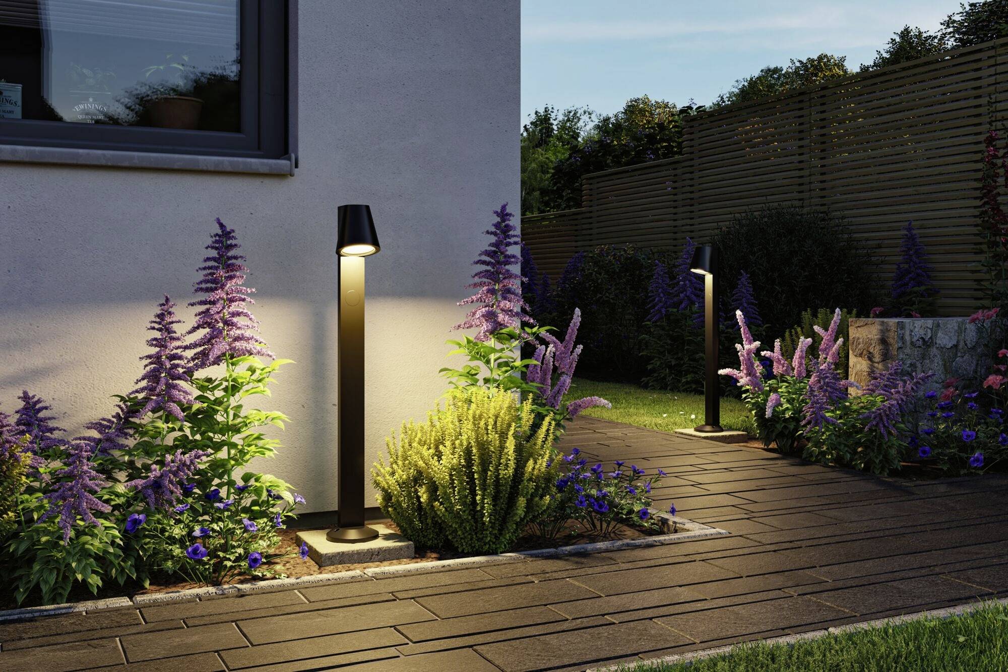 Oświetlona ścieżka ogrodowa z nowoczesnymi latarniami, otoczona fioletowymi i zielonymi roślinami, obok domu o zmierzchu.