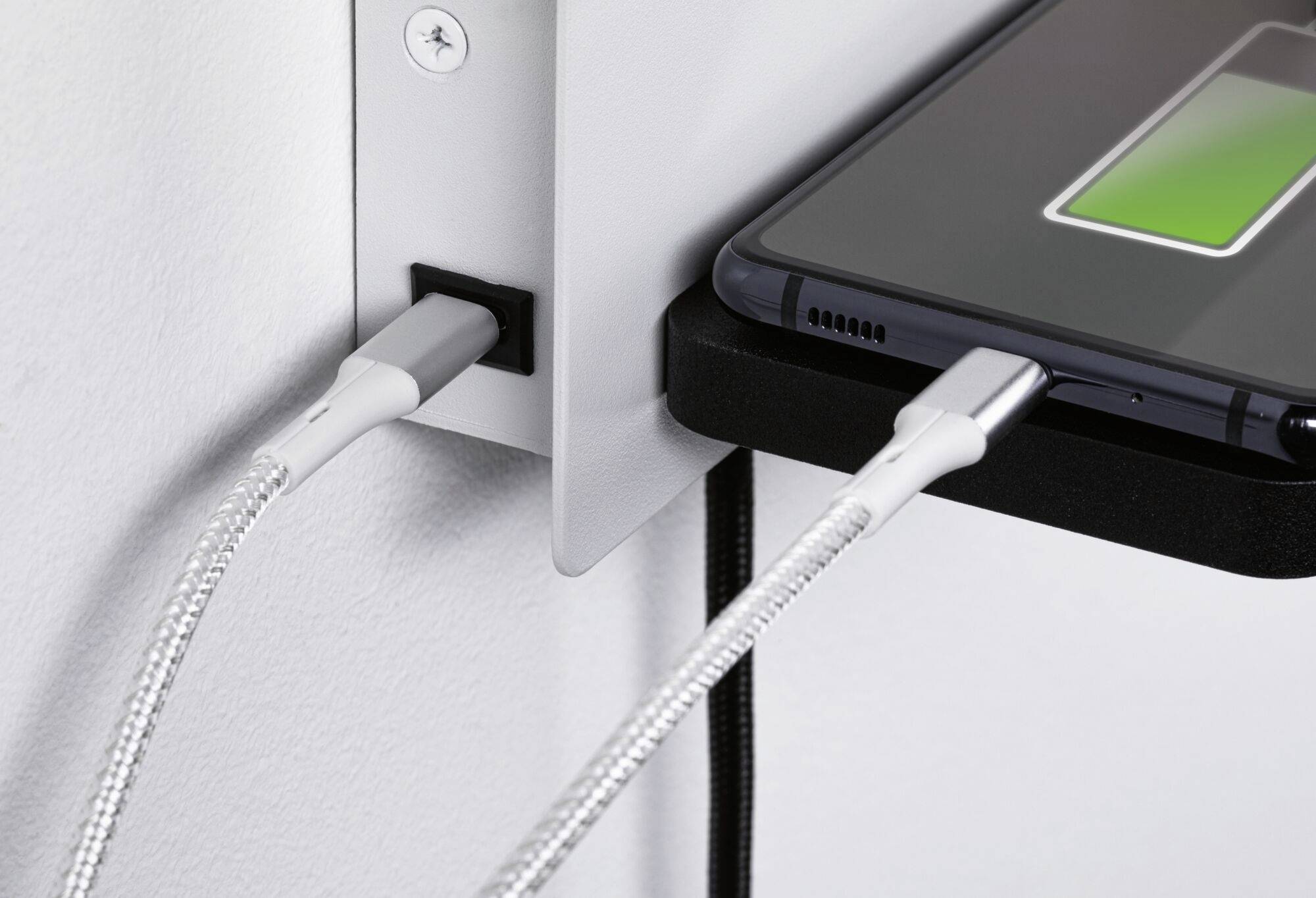 Smartfon ładowany przy ścianie; dwa białe kable USB podłączone do czarnego gniazdka elektrycznego; smartfon wyświetla status ładowania.