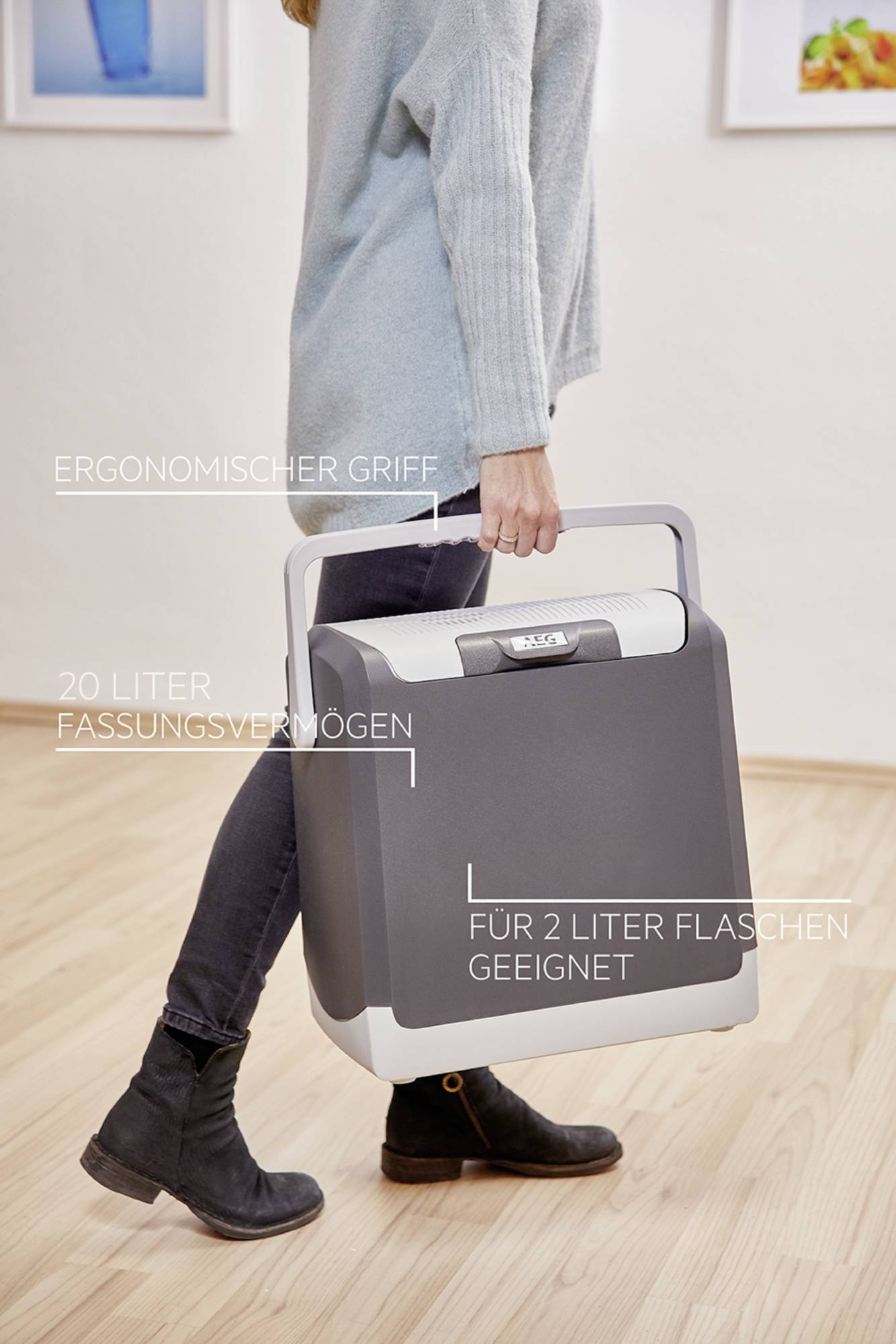 Kobieta niesie przenośną lodówkę turystyczną o pojemności 20 litrów z ergonomicznym uchwytem, mieszczącą butelki 2-litrowe, na jasnej drewnianej podłodze.