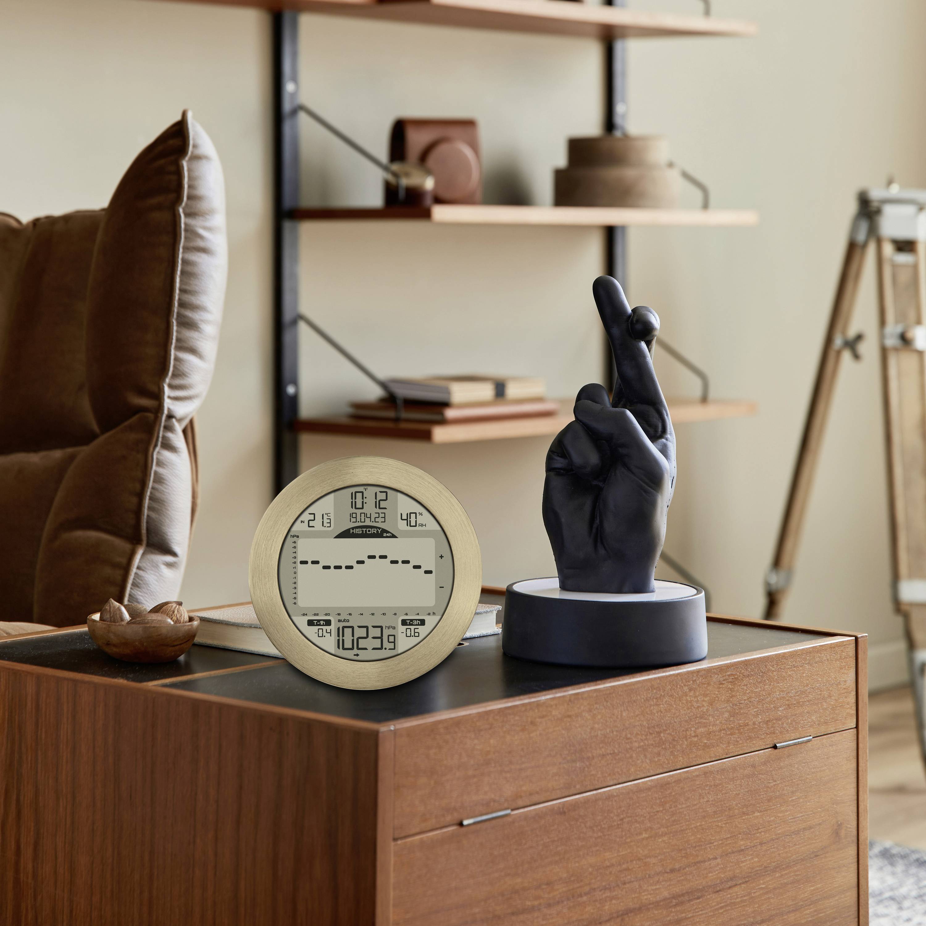 Beżowy cyfrowy budzik z wyświetlaczem czasu i temperatury stoi na drewnianym meblu obok rzeźby dłoni wykonującej gest pokoju.